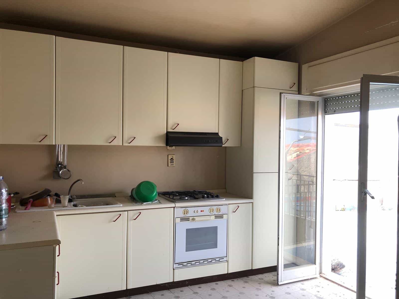 Cucina abitabile con armadi bianchi e forno a gas, finestra con balcone e luce naturale, spazi funzionali e arredo semplice, ideale per appartamenti in affitto o vendita.