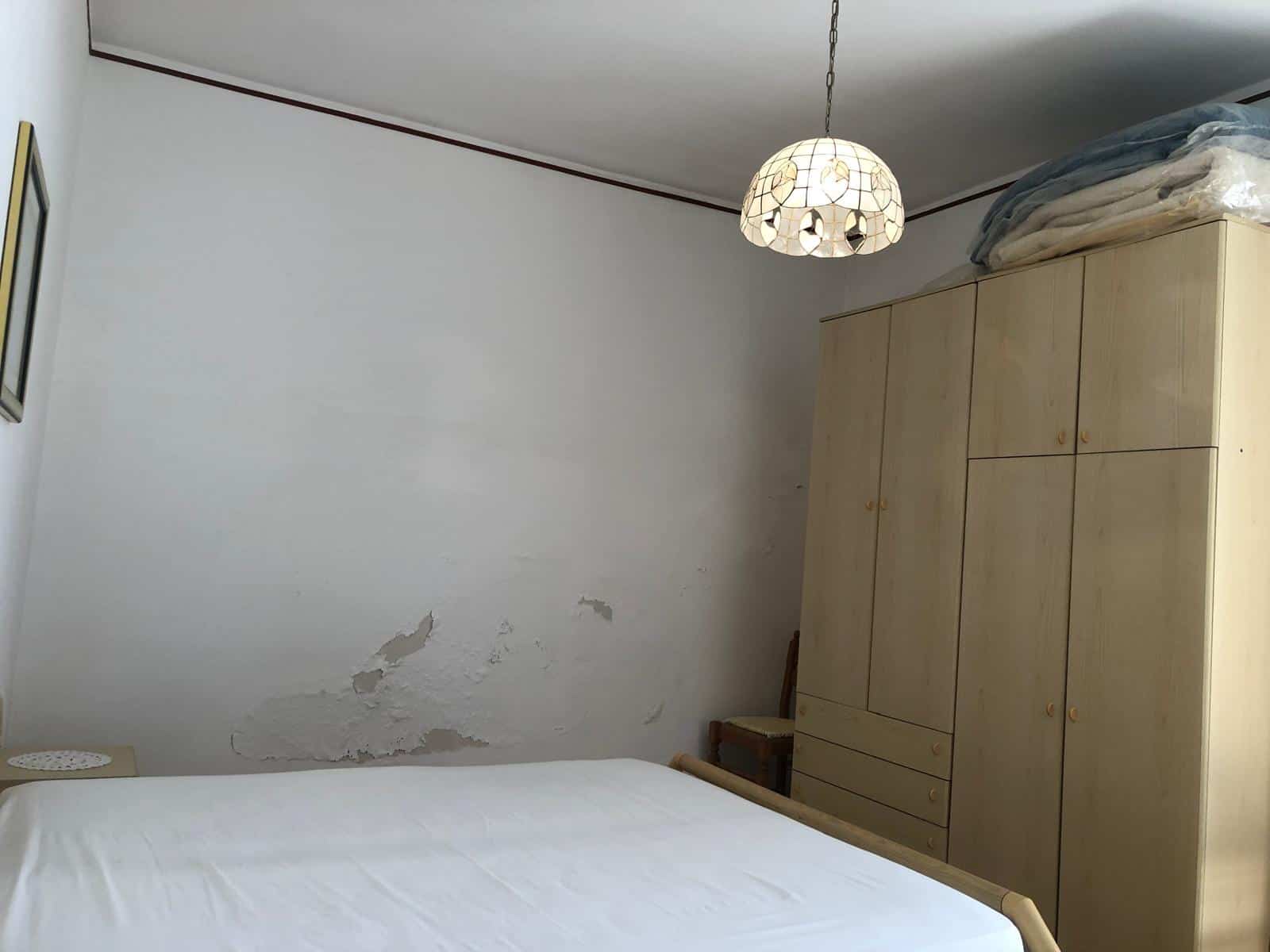 Vecchio armadio in legno chiaro con ripiani e cassetti, letto matrimoniale con lenzuola bianche, parete con segni di umidità e peeling, lampada a soffitto con design decorativo, ambiente rustico e semplice.