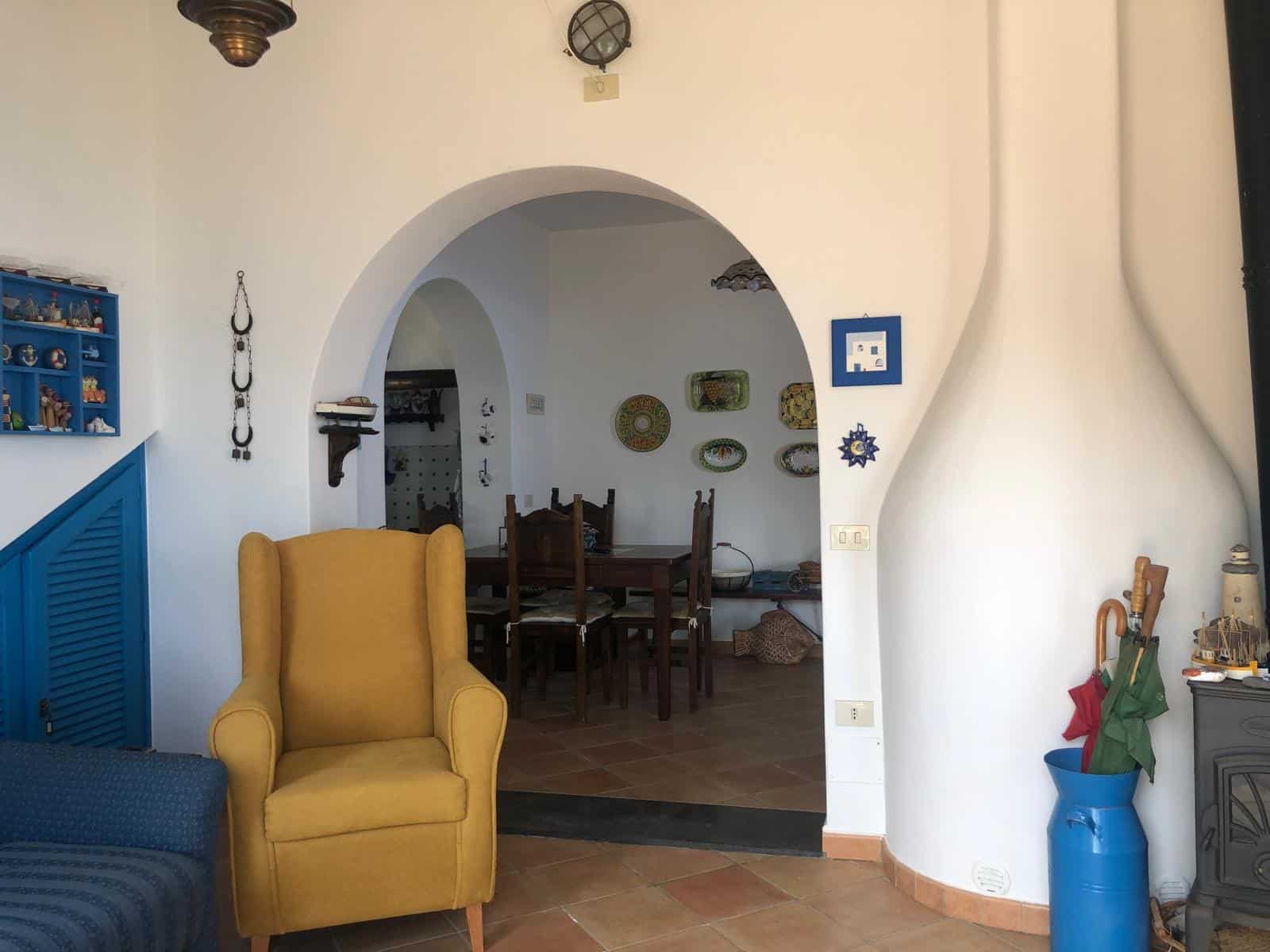 Soggiorno in stile tradizionale con arredi colorati, pareti bianche, archi decorativi, angolo con scaffale blu e poltrona gialla, atmosfera accogliente e originale.