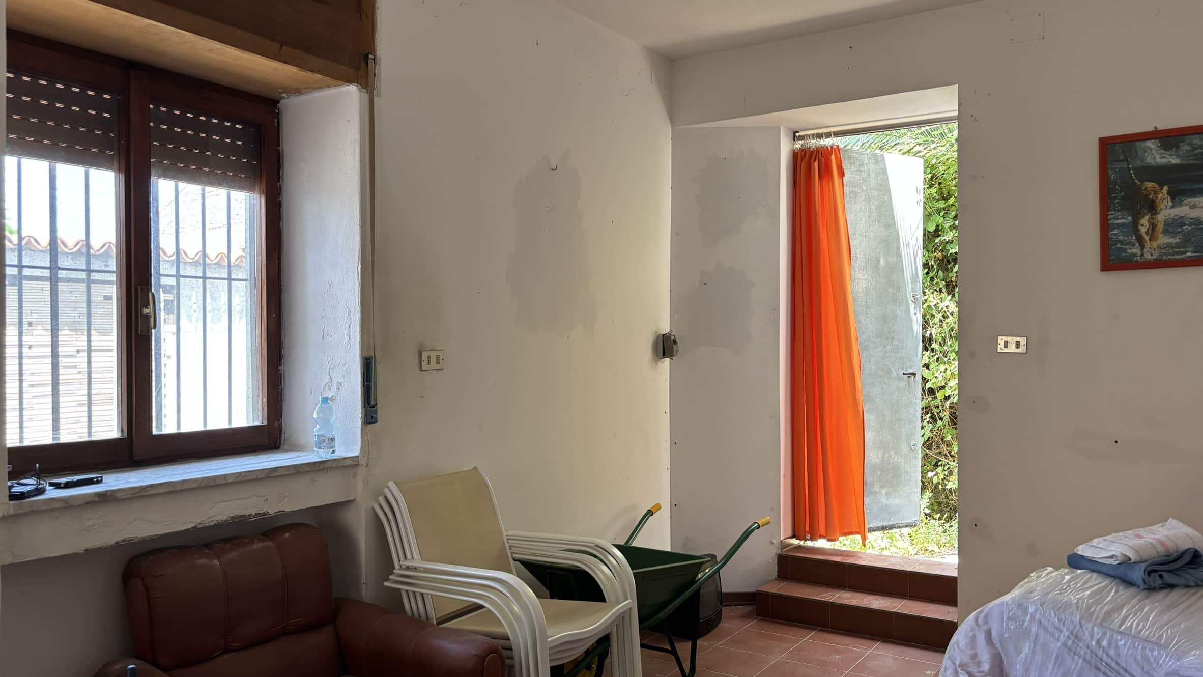 Finestra con inferriata e porte d'ingresso con tenda arancione in una stanza di casa in stile rustico, con mobili impilati e pareti semplici, luce naturale, ambiente accogliente, casa vacanza, ristrutturazione.