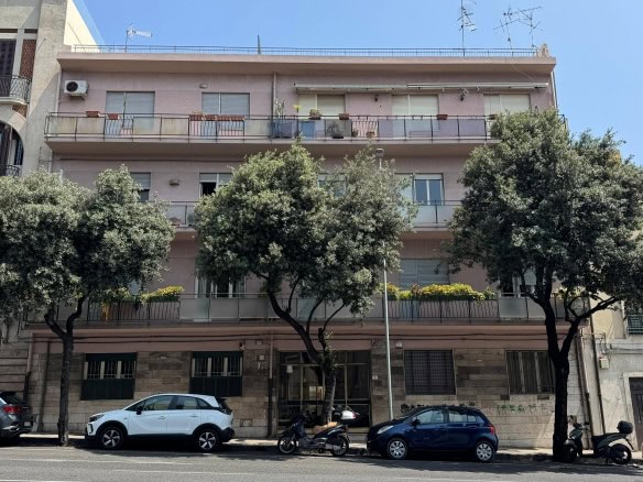 Viale di fronte a un edificio residenziale con balconi, alberi e auto parcheggiate, mostrando un'architettura urbana moderna e alberature che creano un ambiente accogliente e vivace in città.