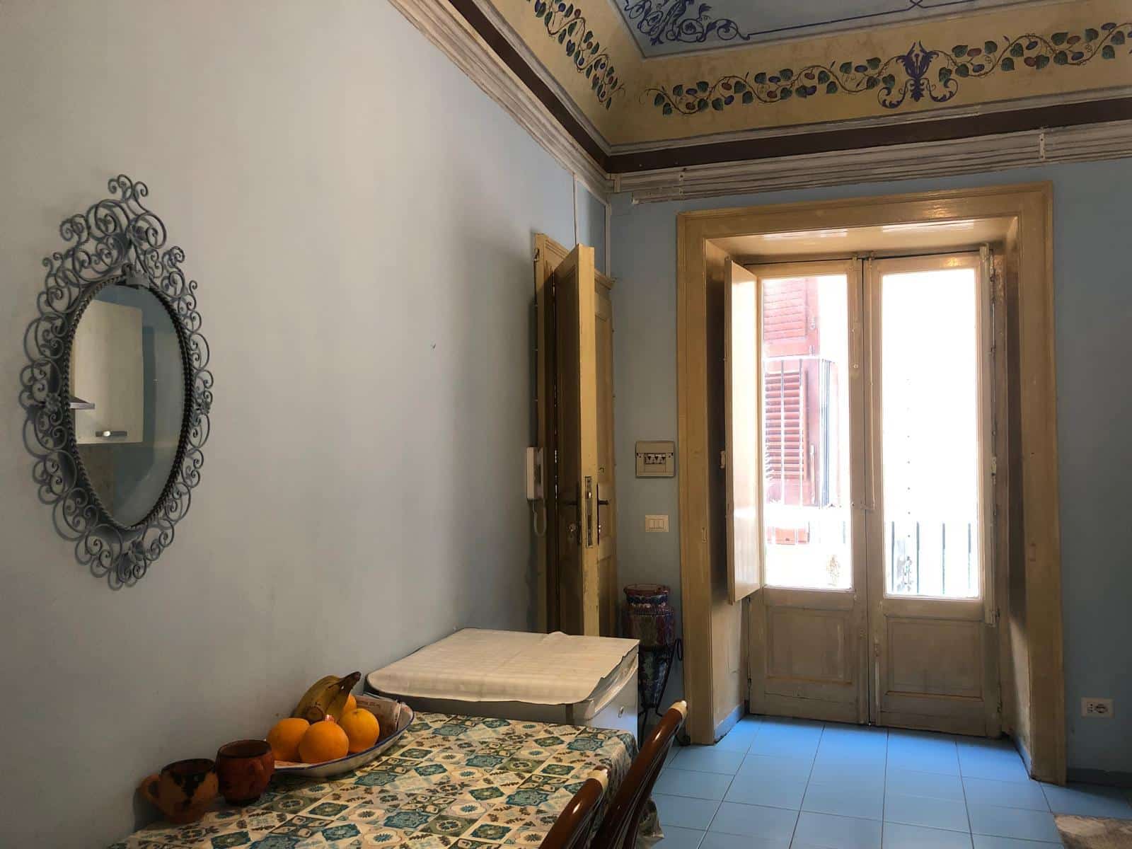 Vecchia porta finestra in legno con persiane in stile classico, illuminata dalla luce naturale, in un soggiorno abitabile con pareti dipinte in azzurro, arredato con un tavolo con tovaglia decorata e specchio in ferro battuto.