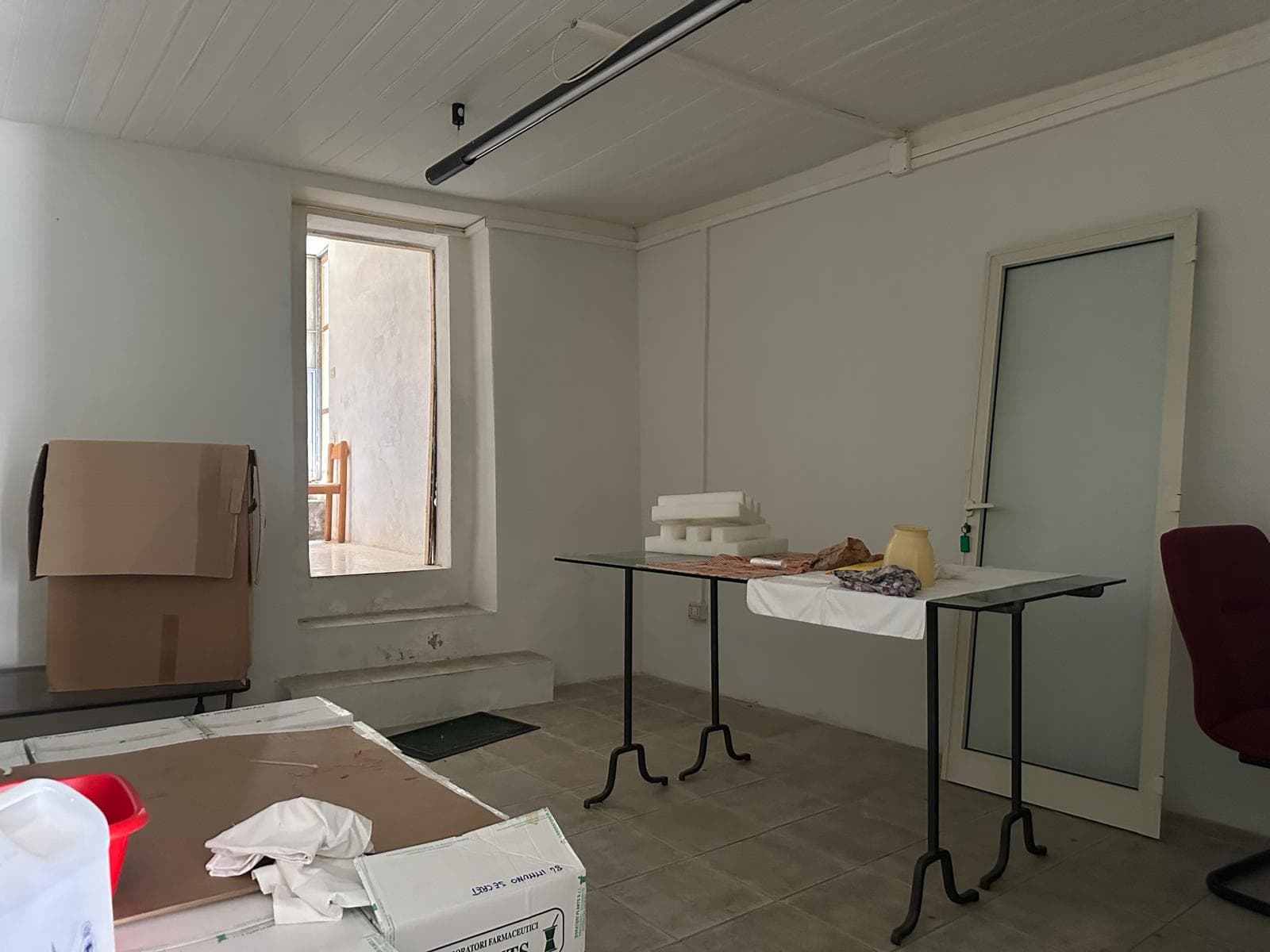 Arredamento ufficio in fase di allestimento con scrivanie e materiali di consumo, parete bianca, porta scorrevole in vetro, finestra aperta che mostra un balcone, atmosfera di lavoro in costruzione.