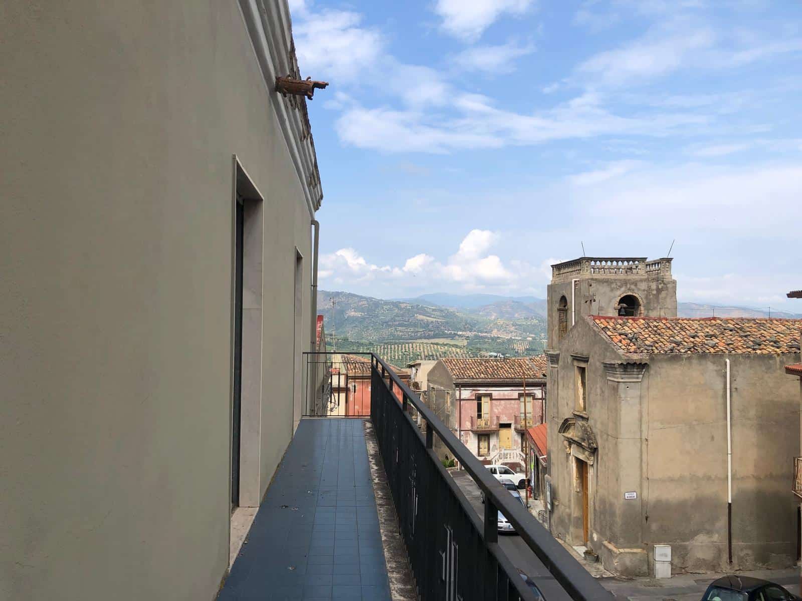 Balcone panoramico con vista sulla città e le colline toscane, edificio storico in stile rustico, tetto in terracotta, giornata soleggiata con cielo azzurro e nuvole bianche, ambiente tranquillo e suggestivo.
