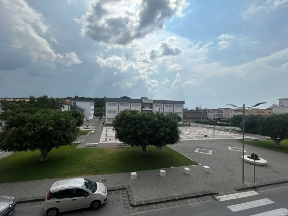Spazio urbano con piazza di interesse pubblico, alberi verdi e parcheggi, vista dalla finestra di un appartamento, con cielo nuvoloso e ambientazione moderna.