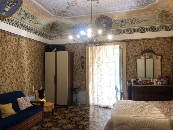 Camere da letto storiche con soffitto decorato, grande armadio e mobili vintage, fantasia floreale alle pareti, lampadario a braccio e balcone con tende leggere, perfette per appartamenti di charme.