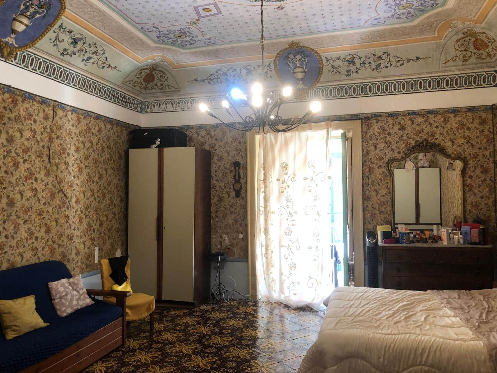 6f79a973-7ae1-43f6-b07b-d1711a666cc2 Camere da letto storiche con soffitto decorato, grande armadio e mobili vintage, fantasia floreale alle pareti, lampadario a braccio e balcone con tende leggere, perfette per appartamenti di charme.