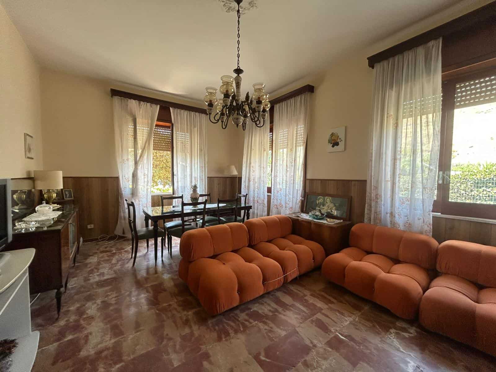 Ampio soggiorno con divani vintage arancioni, illuminato da grandi finestre con tende leggere, arredato con mobili classici e dettagli russi. Ambiente accogliente e luminoso, ideale per relax e convivialità.
