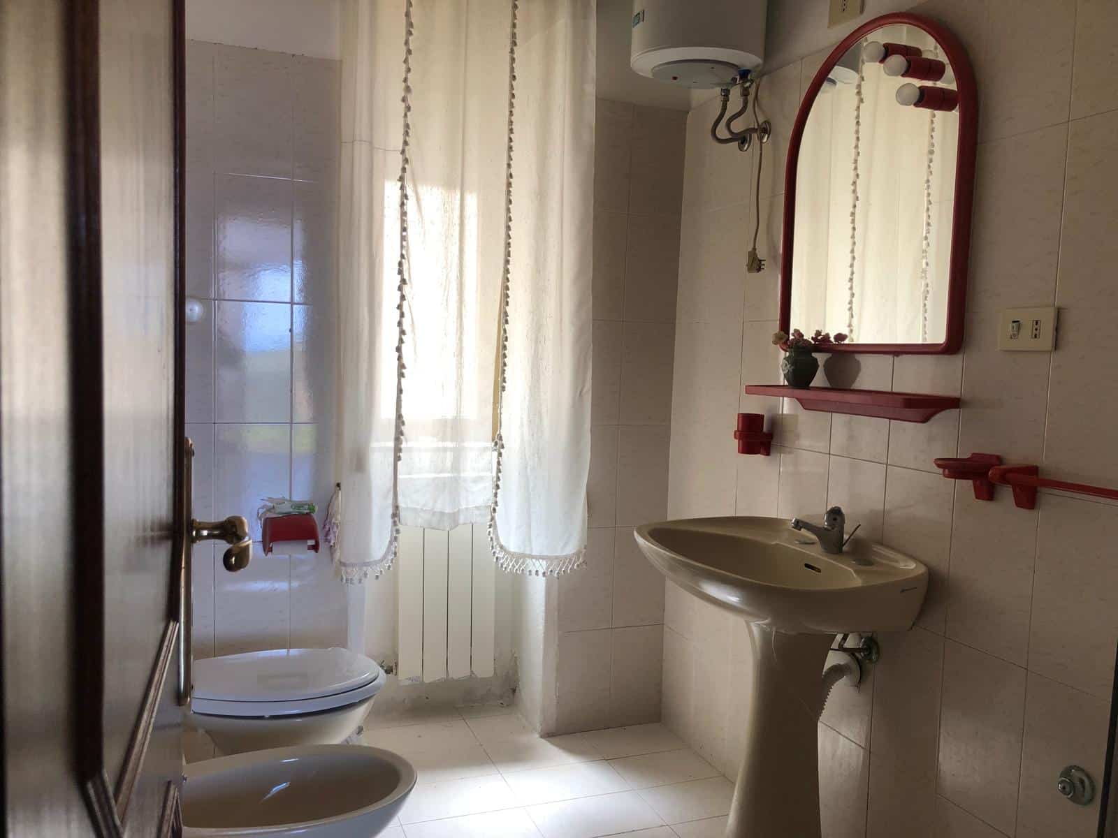 Comforte bagno con lavabo, specchio e arredamento rosa, finestra con tende leggere, servizi igiene e ambiente ristrutturato, ideale per affitti o uso privato.