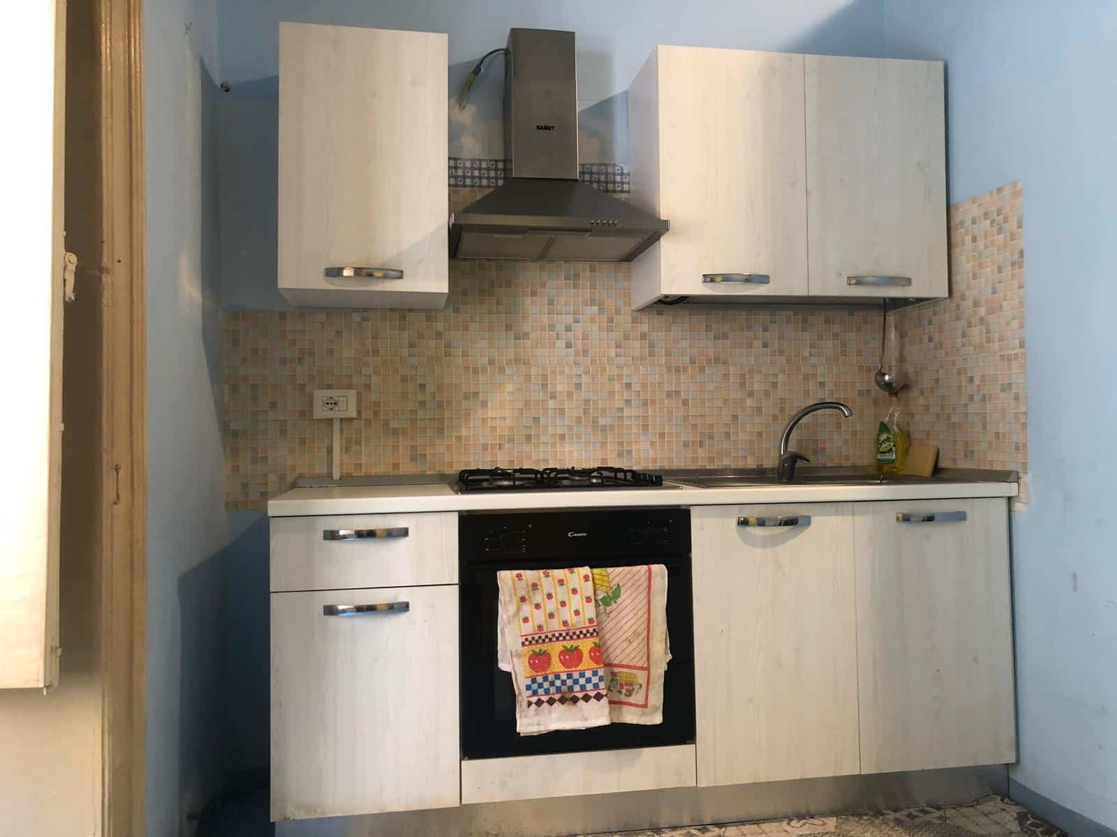 Cucina compatta con ante in stile rustico, piano cottura a gas e zona lavabo, ideale per spazi piccoli e funzionali. Caratterizzata da pareti rivestite in mosaico beige, arredi essenziali e pratici.