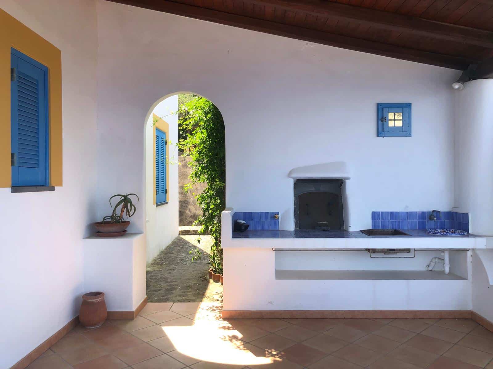 Finestra con persiana blu e cucina esterna in stile mediterraneo, pareti bianche e dettagli colorati, ambiente accogliente e soleggiato, ideale per vivere la tradizione e il relax all'aperto.