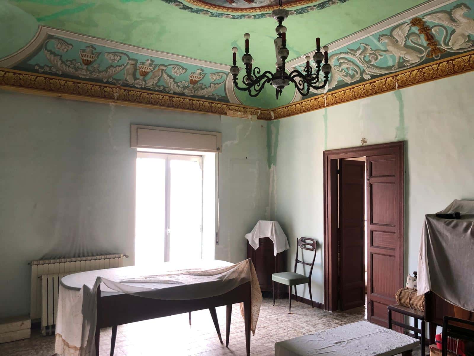 Antico ambiente interno con soffitto decorato e lampadario d'epoca, finestra luminosa, mobili classici e pareti con tracce di ristrutturazione, arredamento vintage, stile storico e rustico, NextCasa Italia.