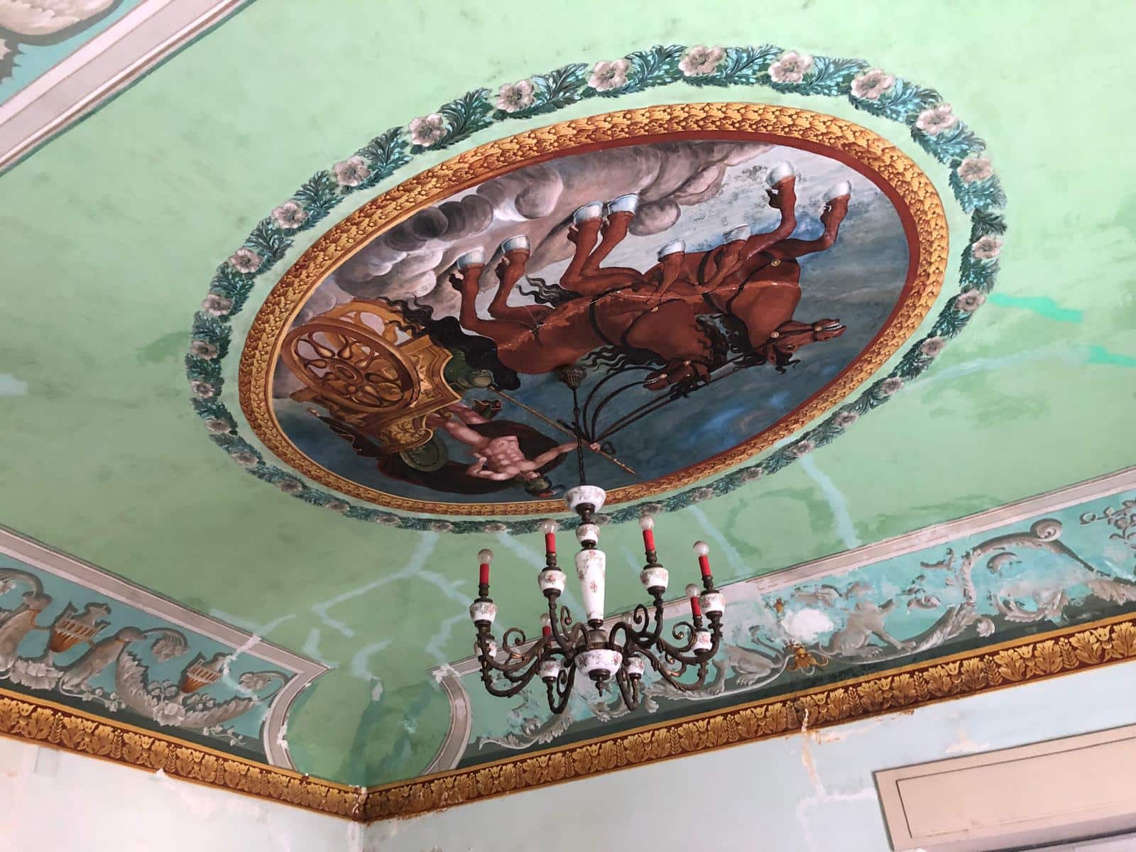 Trompe-l'œil artistico sul soffitto con dipinto storico raffigurante cavalli e carrozza, ornato da dettagli decorativi dorati e una lampadaria antica, creando un'atmosfera elegante e raffinata.