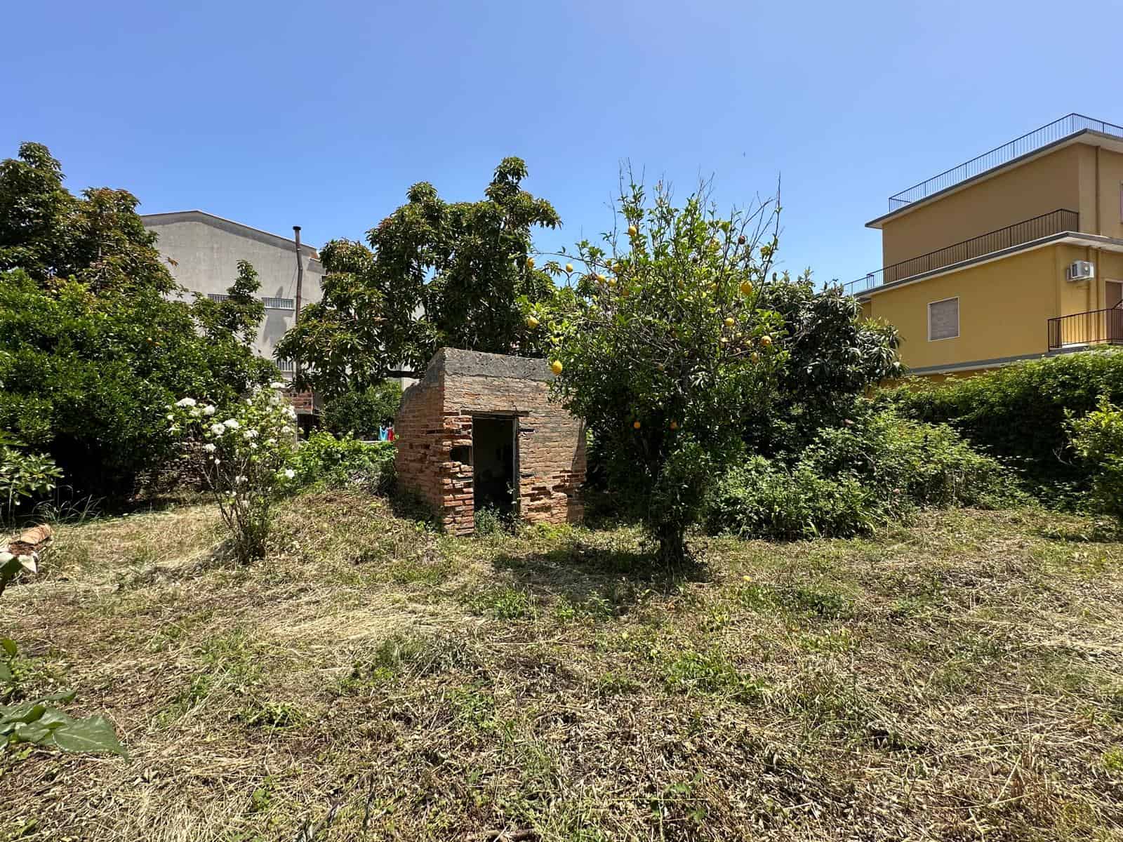 Rudere di campagna ex casetta in mattoni rossi circondata da alberi e piante fruttifere in un giardino urbano con edifici moderni sullo sfondo.