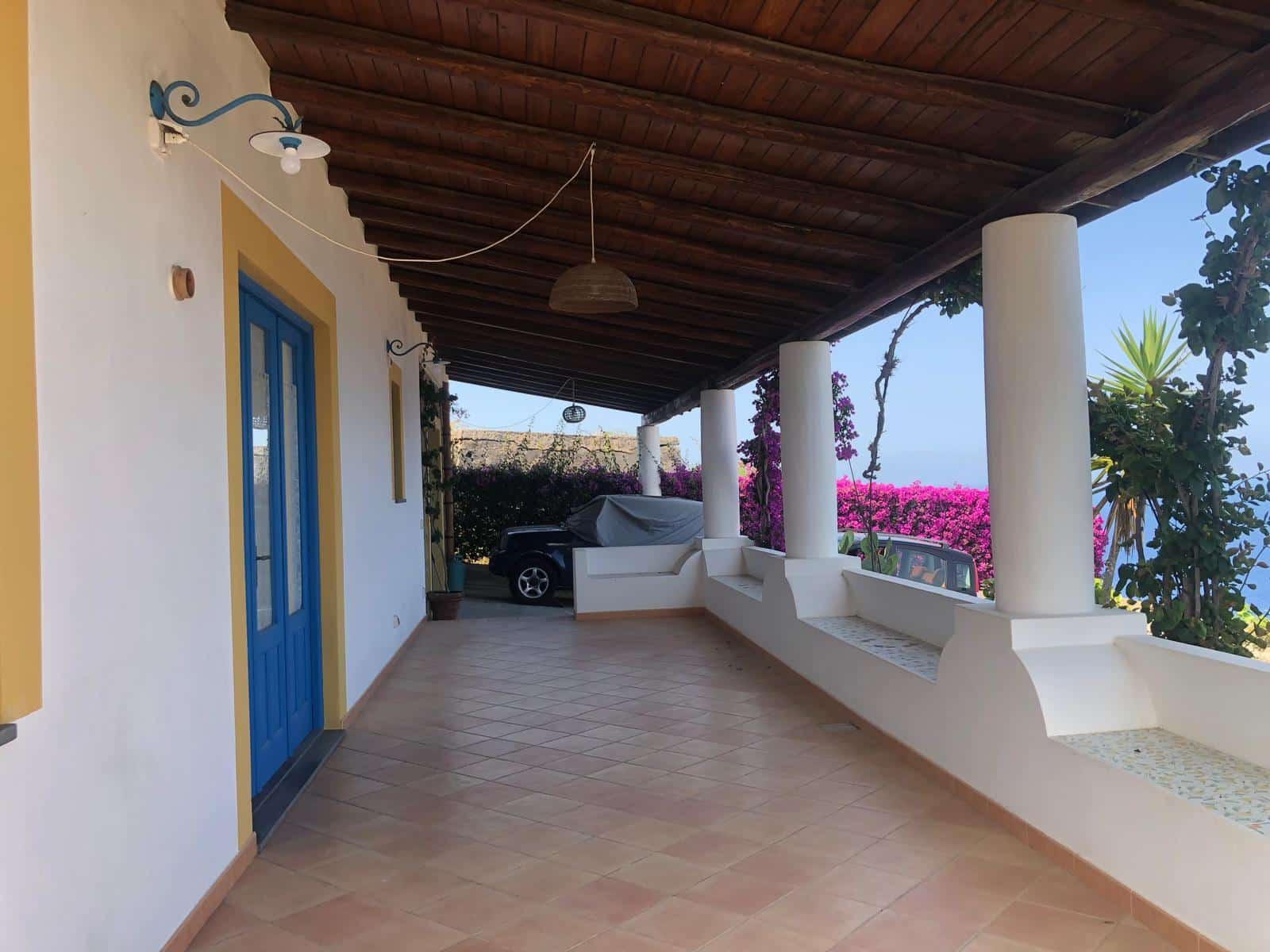 Balcone di una casa in stile mediterraneo con veranda coperta, colonne bianche, porte blu, piante e fiori colorati, vista panoramica mare, atmosfera rilassante e accogliente, ideale per case vacanza NextCasa.
