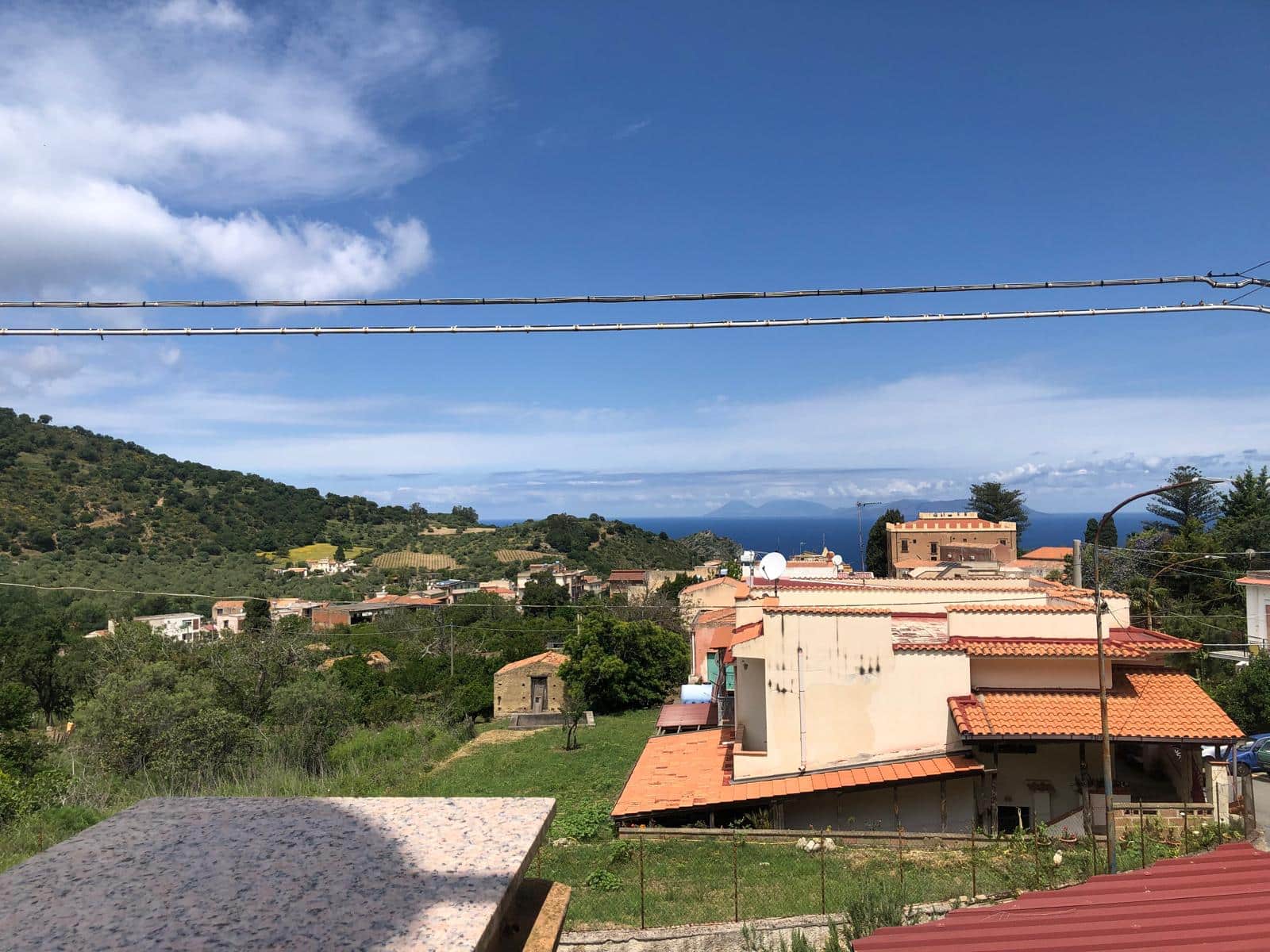 Veduta panoramica delle case del villaggio con tettori in ceramica, colline verdi e mare in lontananza, sotto un cielo sereno e nuvoloso, immagine di esclusiva proprietà e bellezze naturali nel contesto di NextCasa.