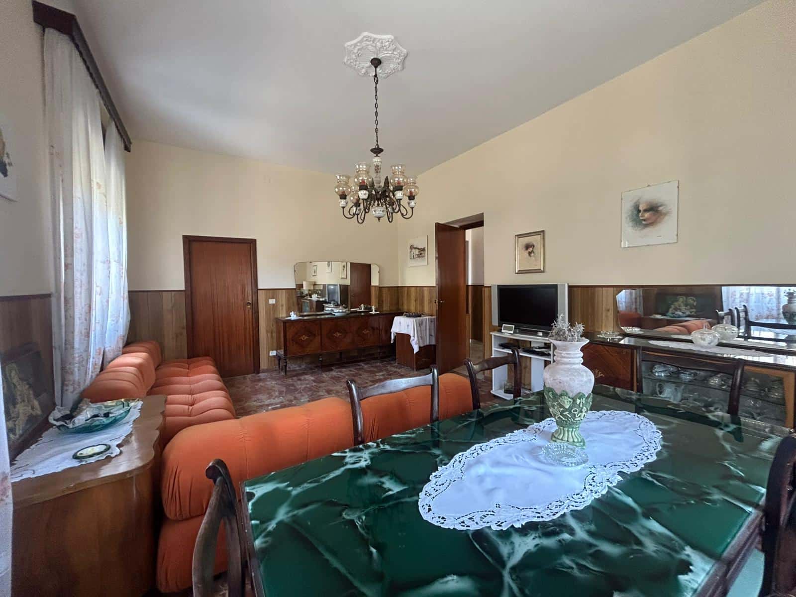 Vecchia sala da pranzo con divani arancioni, tavolo con tovaglia verde, mobili in stile classico e decorazioni vintage, ambiente accogliente e ben arredato, ideale per vivere momenti in famiglia.