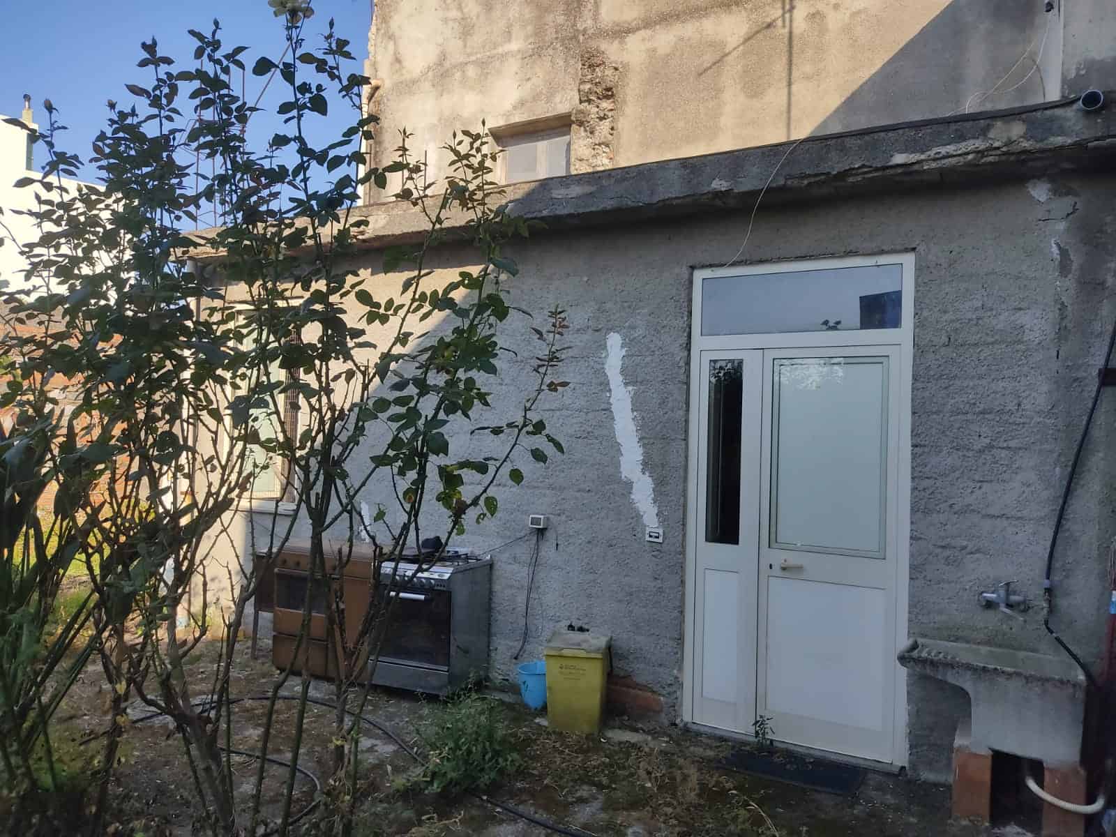 Vecchia casa con cortile esterno, porta in PVC bianco, finestra superiore, area esterna con piante e giardino, forno da esterno e utilità per il giardinaggio, ristrutturazione e rinnovamento edilizio, immobile abitativo rustico.