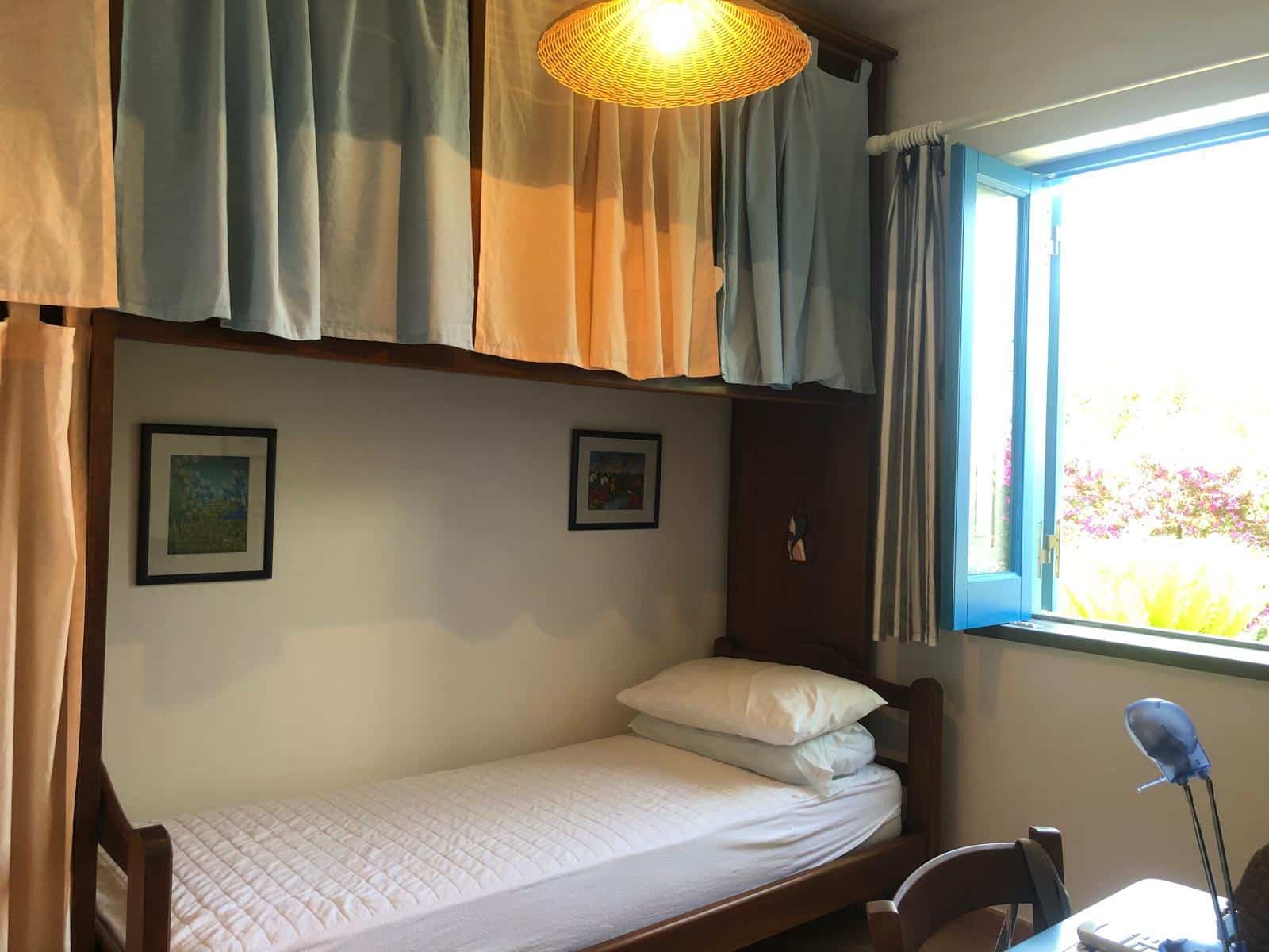 Letto singolo con cuscini in una camera luminosa con finestra aperta, decorazioni floreali e tende colorate, atmosfera accogliente e rilassante, ideale per soggiorni brevi o in affitto, NextCasa.