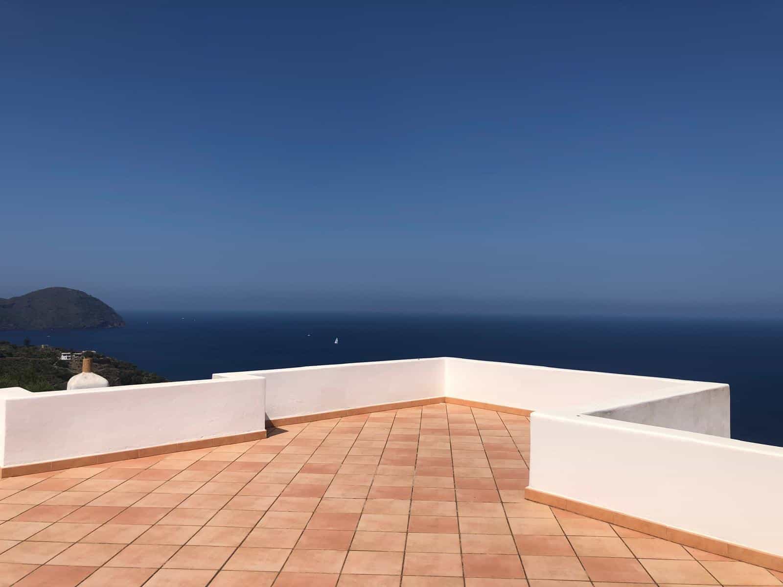 Spiaggia vista mare da un terrazzo con parapetto bianco e pavimento in piastrelle terracotta, aspetto rilassante e panoramico, ideale per case vacanza o appartamenti di lusso a NextCasa.