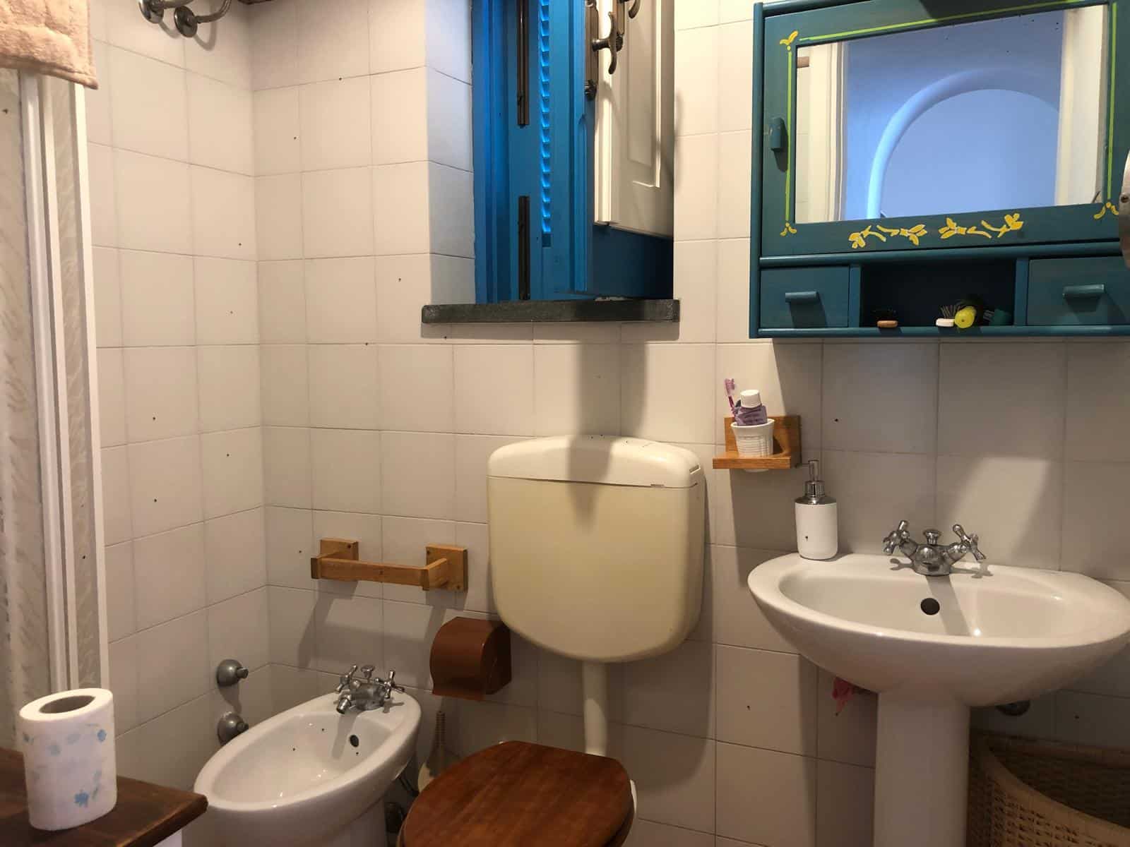 Bagno di casa rustico con lavabo, bidet e specchio azzurro, arredamento in stile tradizionale, piastrelle bianche, dettagli in legno, ambiente accogliente e funzionale sotto luce naturale.