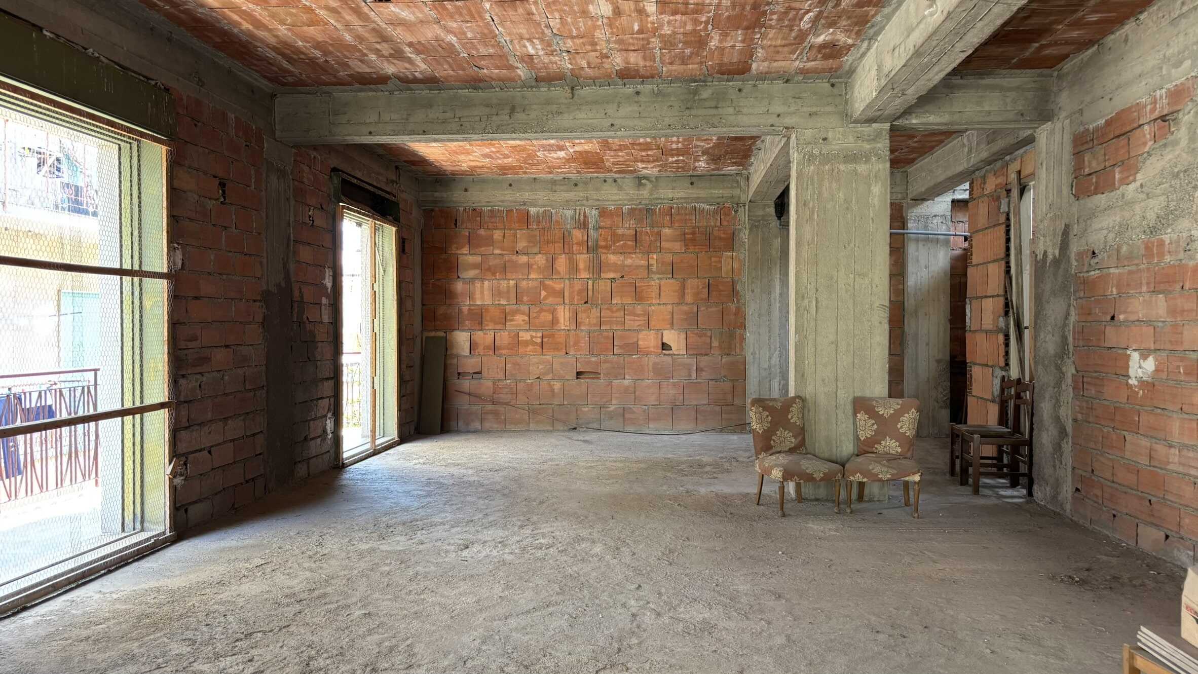 Angolo di un appartamento in costruzione con pareti in mattoni e struttura in cemento, grandi finestre per illuminazione naturale, con alcuni mobili antichi e caratteristiche di open space.