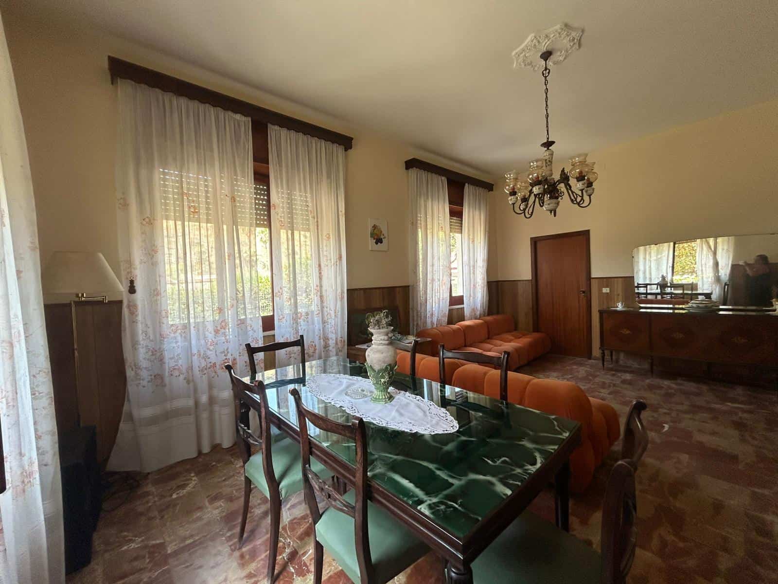 Spazio soggiorno con mobili vintage, divano arancione e tavolo in marmo verde, atmosfera calda e accogliente, stile retrò, grande finestra con tende floreali, illuminazione soffusa, arredi classici, atmosfera di casa famigliare.