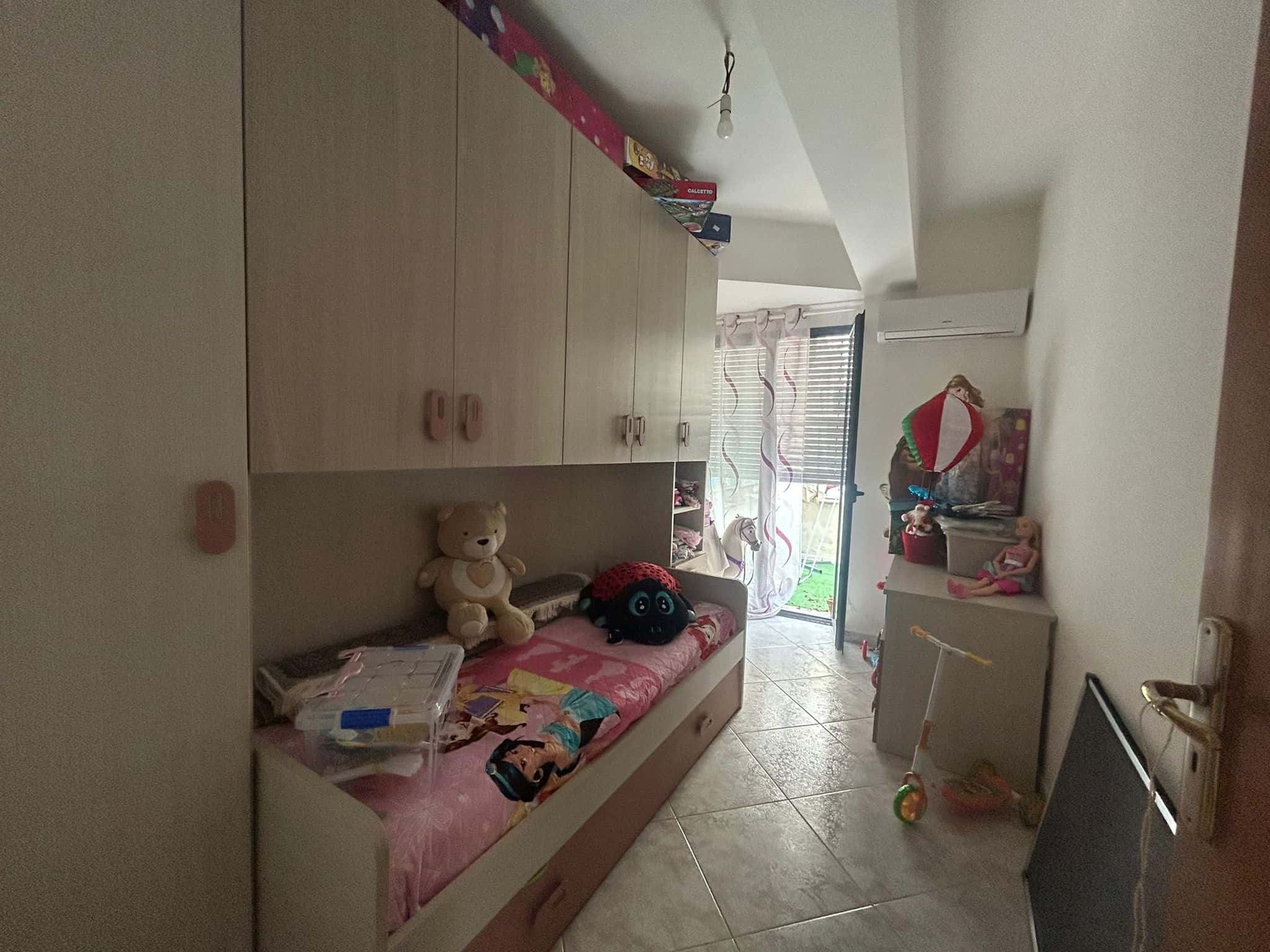 Letto singolo con armadio a ponte, cameretta per bambini con giochi e giocattoli, spazio accogliente e funzionale, ambiente luminoso con accesso al balcone, atmosfera casalinga e ordinata.