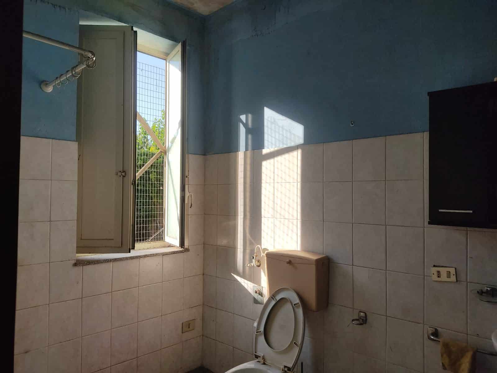 Finestra aperta con vista su giardino in bagno con pareti blu e piastrelle beige, luce naturale e stile semplice, ideale per ristrutturazioni e immobili residenziali.