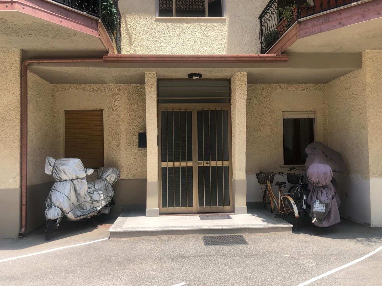 Moto coperta e biciclette sotto l'ingresso di un appartamento in un condominio, con un portone girevole e una finestra. Maestria nel design e sicurezza per la zona abitativa.