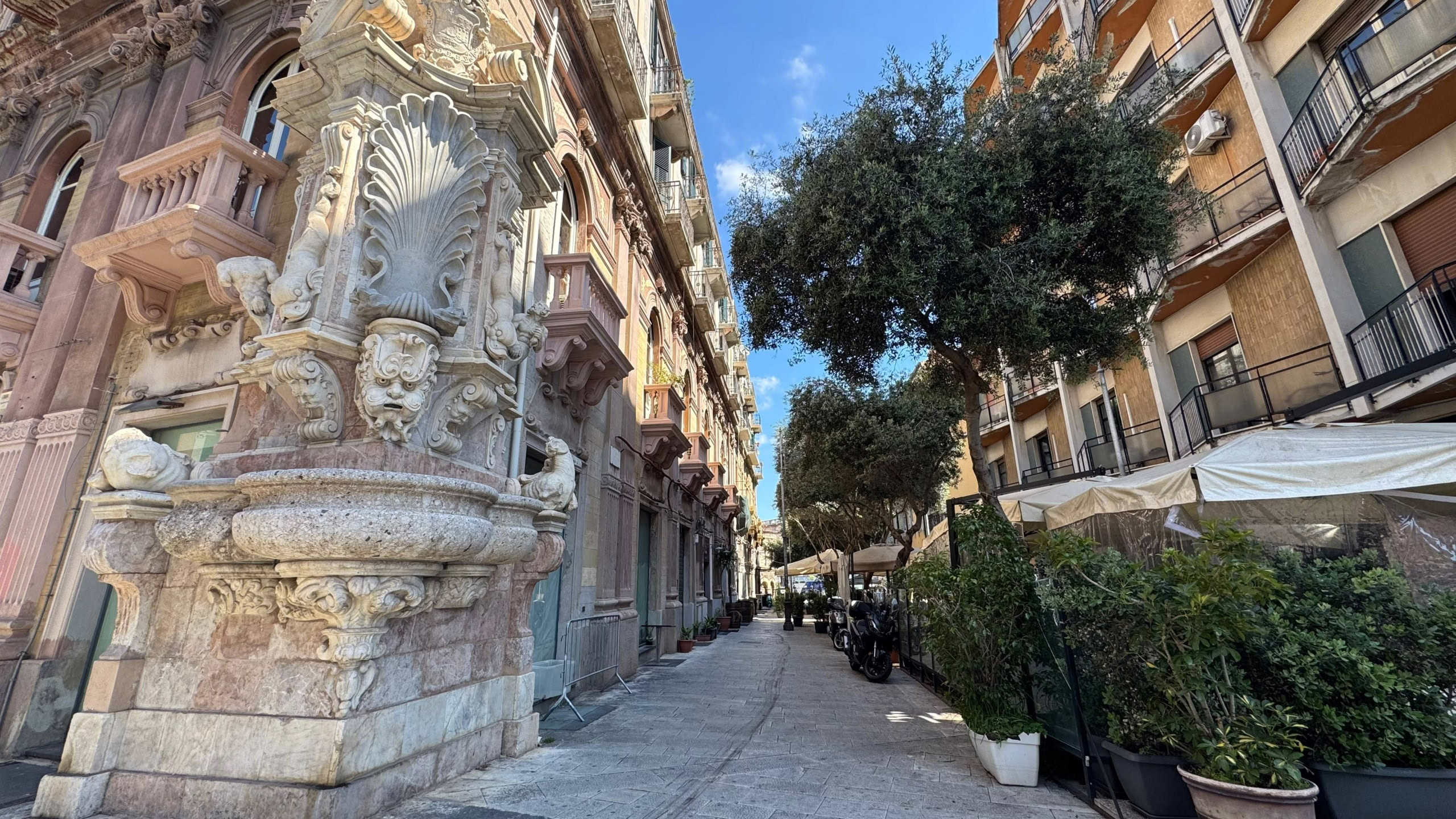 Finestra con decorazioni in stile barocco e fioriere lungo una strada pedonale nel centro di Napoli, Italia, con edifici storici e alberi ombreggiati, rappresentando l’arte e l’architettura classica.