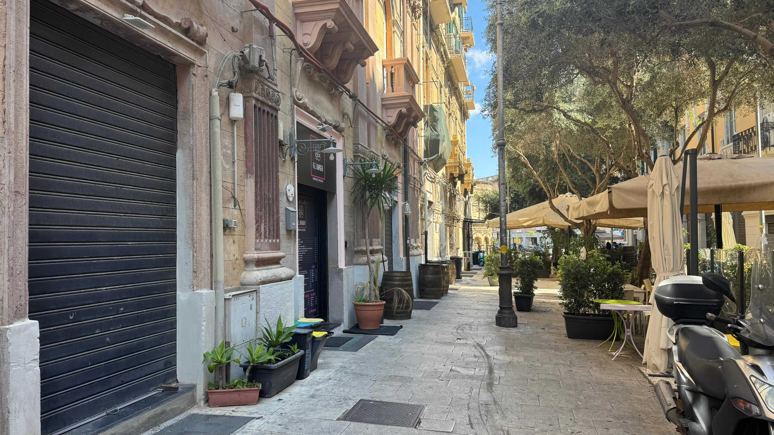 Vicolo cittadino con negozi e caffetterie all'aperto, caratterizzato da edifici storici, piante in vaso, motorette parcheggiate e sedie con ombrelloni per rilassarsi, scenario di un centro urbano mediterraneo.