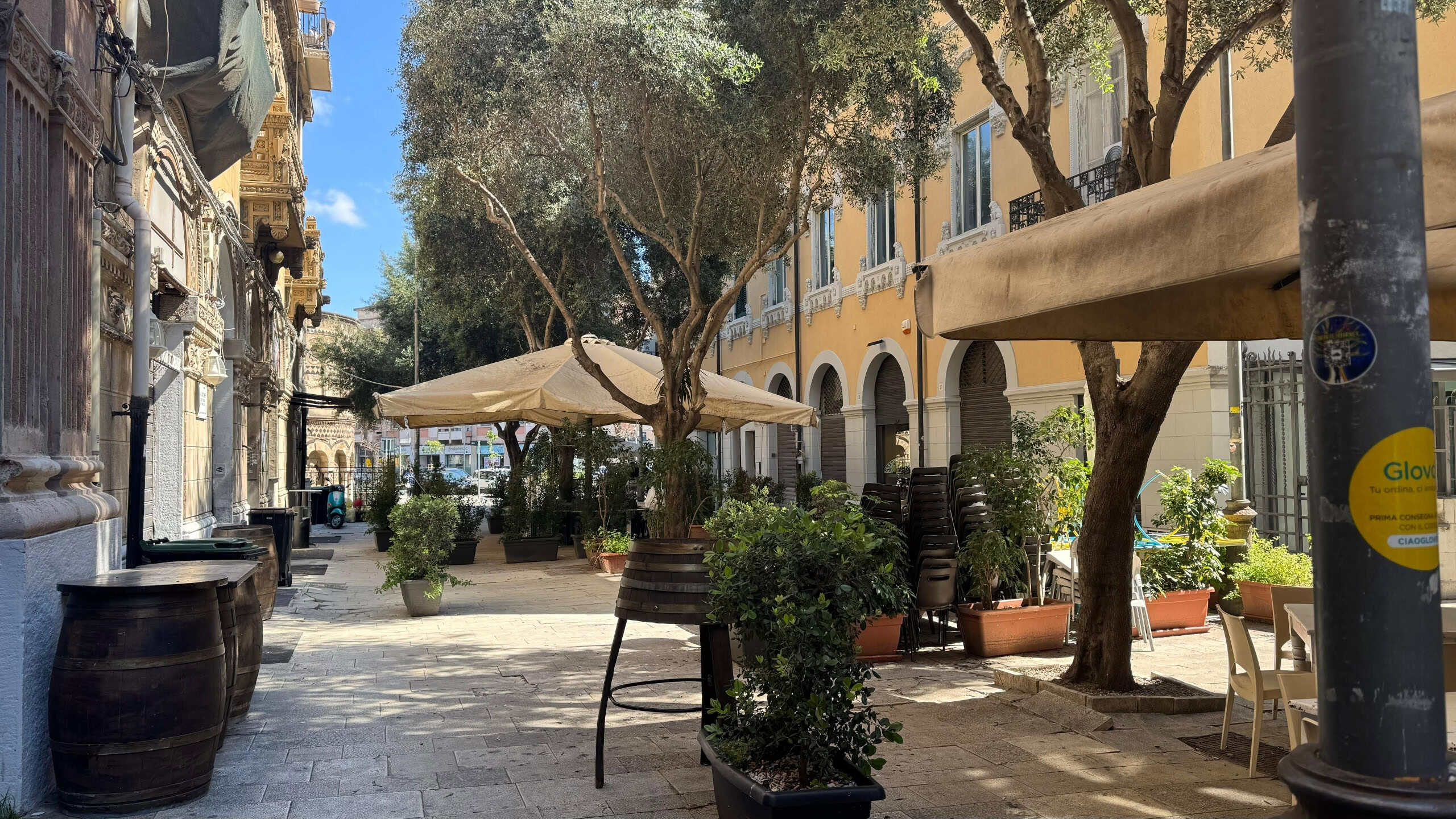 Velia piazza con alberi e sedie all'aperto, atmosfera rilassante, possibile locale di ristorante o caffetteria, elegante edificio in stile classico, Spazio pubblico accogliente e ombreggiato, ideale per socializzare e gustare caffè o pasti all'aperto.