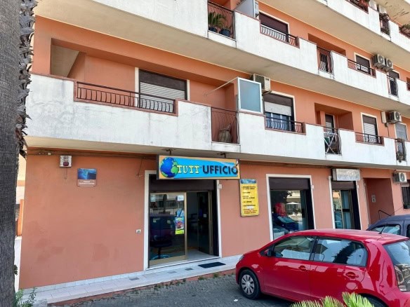 Tutti Ufficio, ufficio commerciale situato al piano terra di un edificio residenziale con facciata color terracotta, con autovettura rossa parcheggiata davanti, segnale commerciale illuminato, in zona urbana.