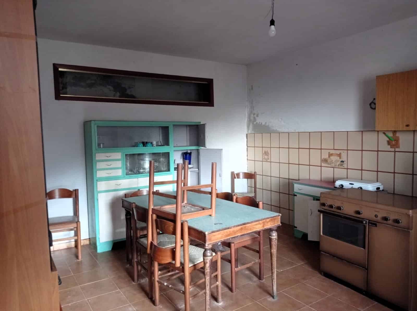 Vecchia cucina rustica con mobili in legno, tavolo da pranzo e forno a gas, pareti con piastrelle e parete bianca, ambiente abitabile da ristrutturare, luce naturale e aria informale.
