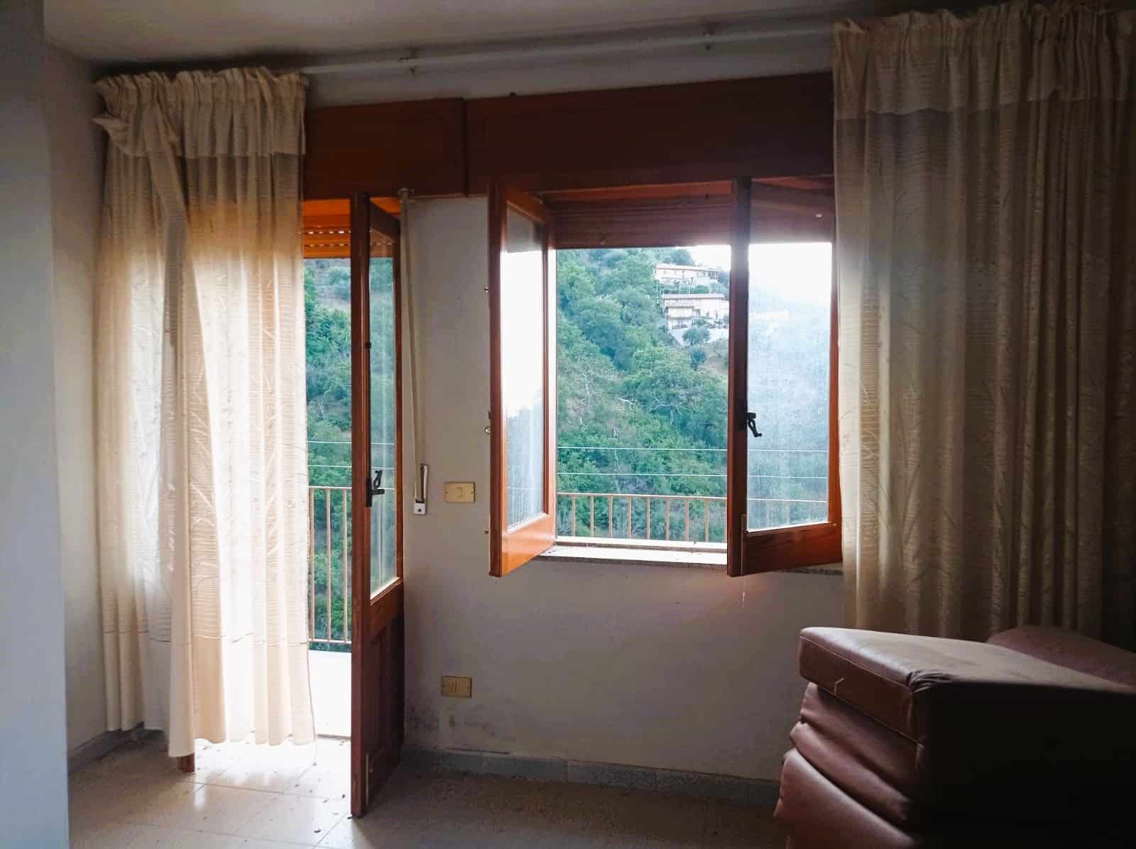 Foto di una stanza con porta finestra e balcone, vista su un paesaggio verde, spazio abitativo luminoso con tende beige, ambiente tranquillo e confortevole.
