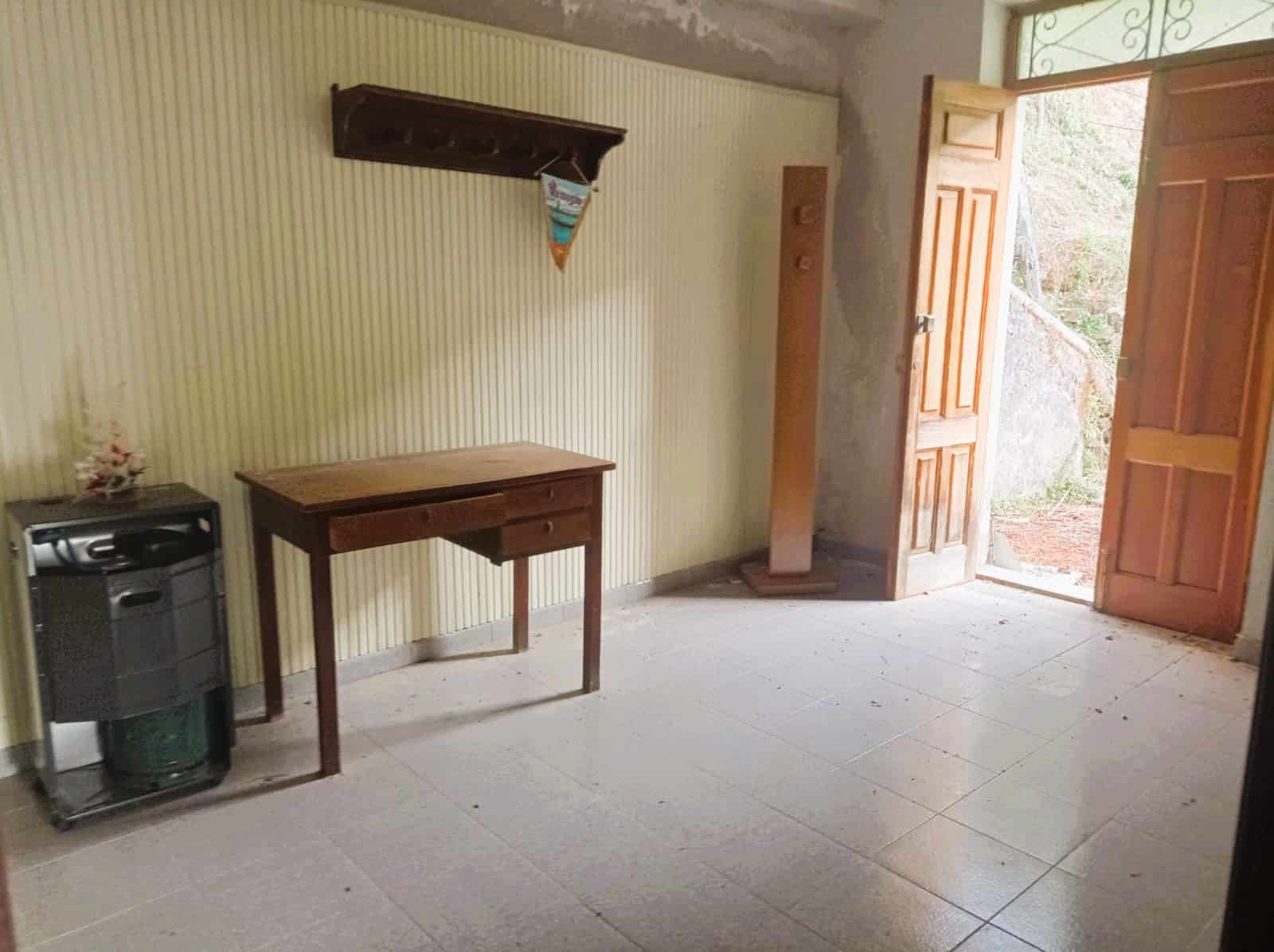 Soggiorno rustico con pareti a strisce e porta in legno aperta che si affaccia su un ambiente esterno naturale. Arredi semplici, tra cui un tavolo in legno e una stufa a pellet, creando un'atmosfera calda e accogliente.
