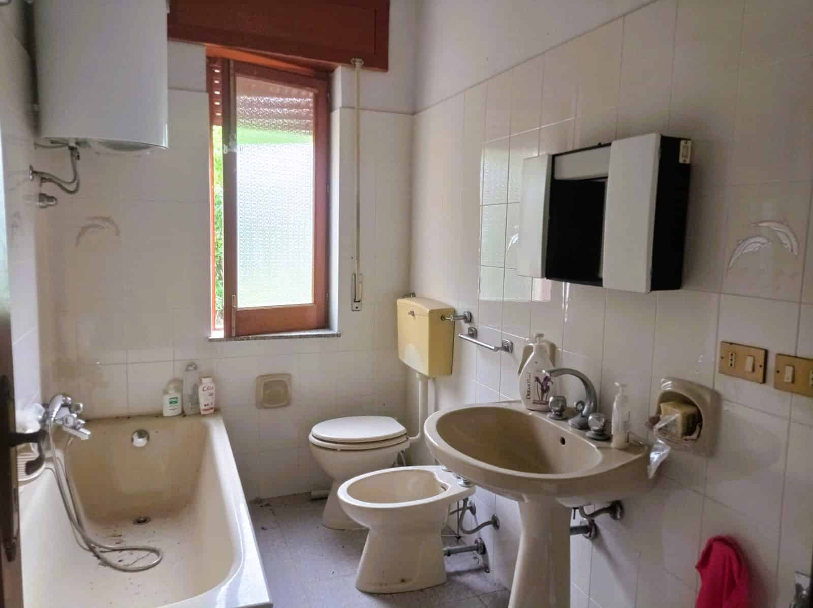 Vecchio bagno con vasca, WC e lavabo in stile vintage, luce naturale dalla finestra in legno, piastrelle chiare, ambiente semplice e funzionale, adatto per ristrutturazione o appartamenti storici.