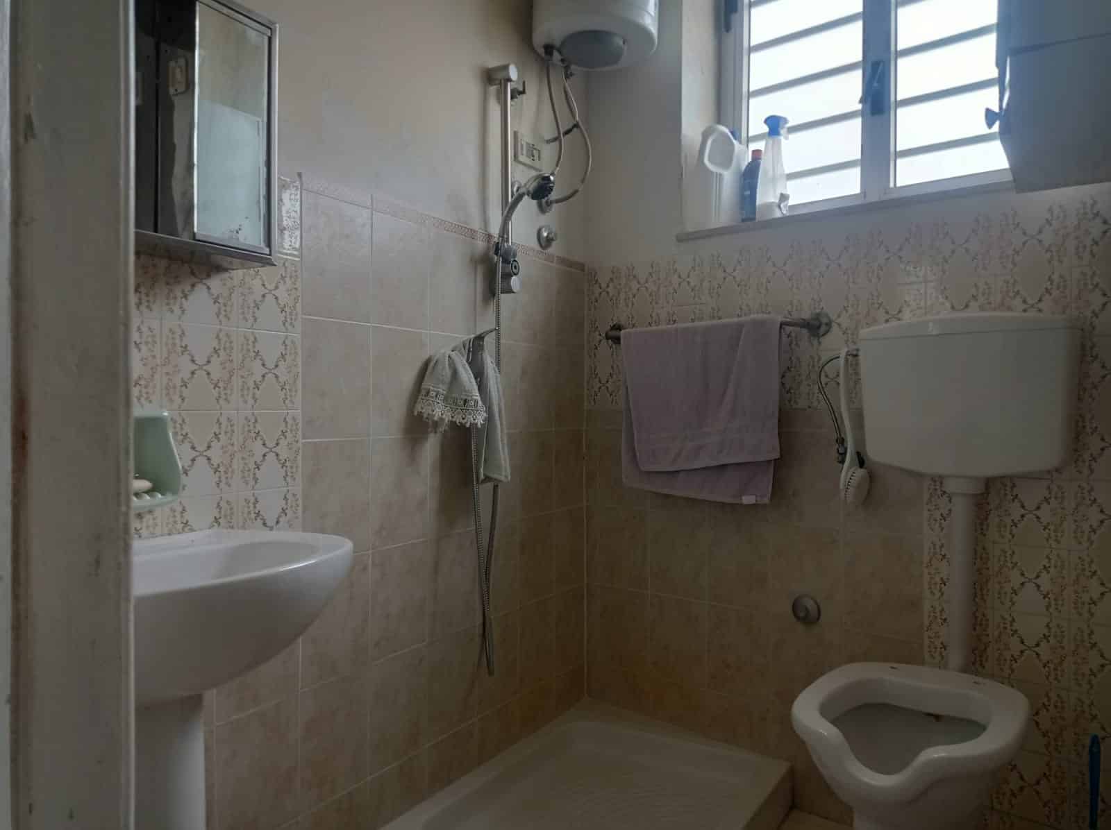 Lavabo, WC e doccia in bagno con finestra ventilata, sanitari tradizionali, pareti rivestite con piastrelle beige con decorazioni, ideale per ristrutturazioni bagno e soluzioni abitative pratiche.