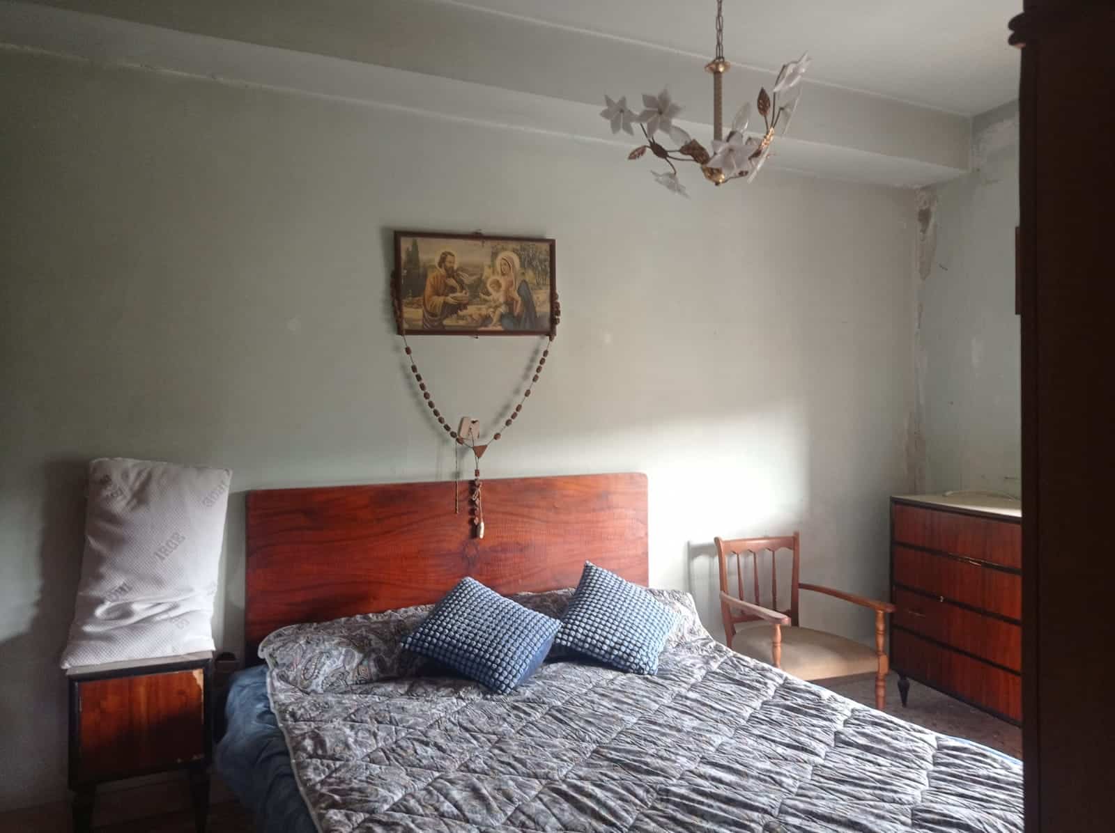Vecchia camera da letto con letto in legno, cuscini a scacchi, soffitto con lampadario e immagine sacra appesa alla parete, mobili vintage, atmosfera rustica e retrò.