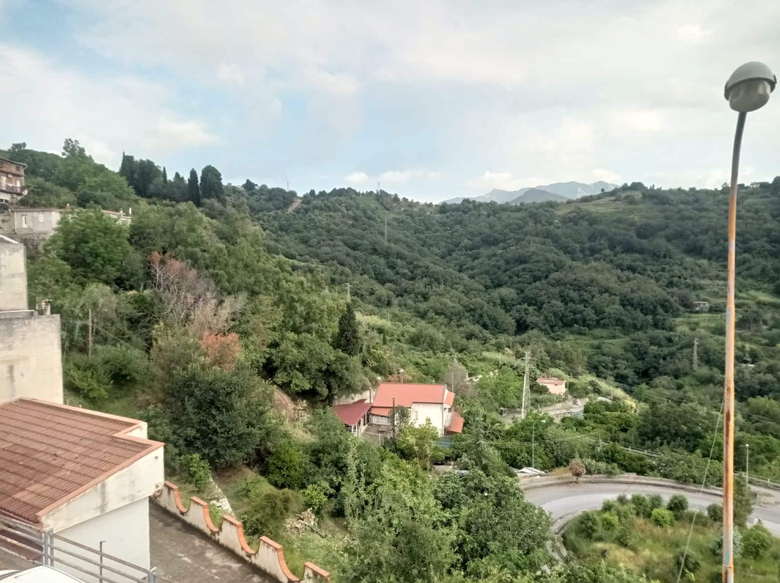 Esterni di case in collina circondate da vegetazione verde, vista panoramica sulle montagne e strade con curve, scena naturale e tranquilla, ideale per real estate e agenzie immobiliari.