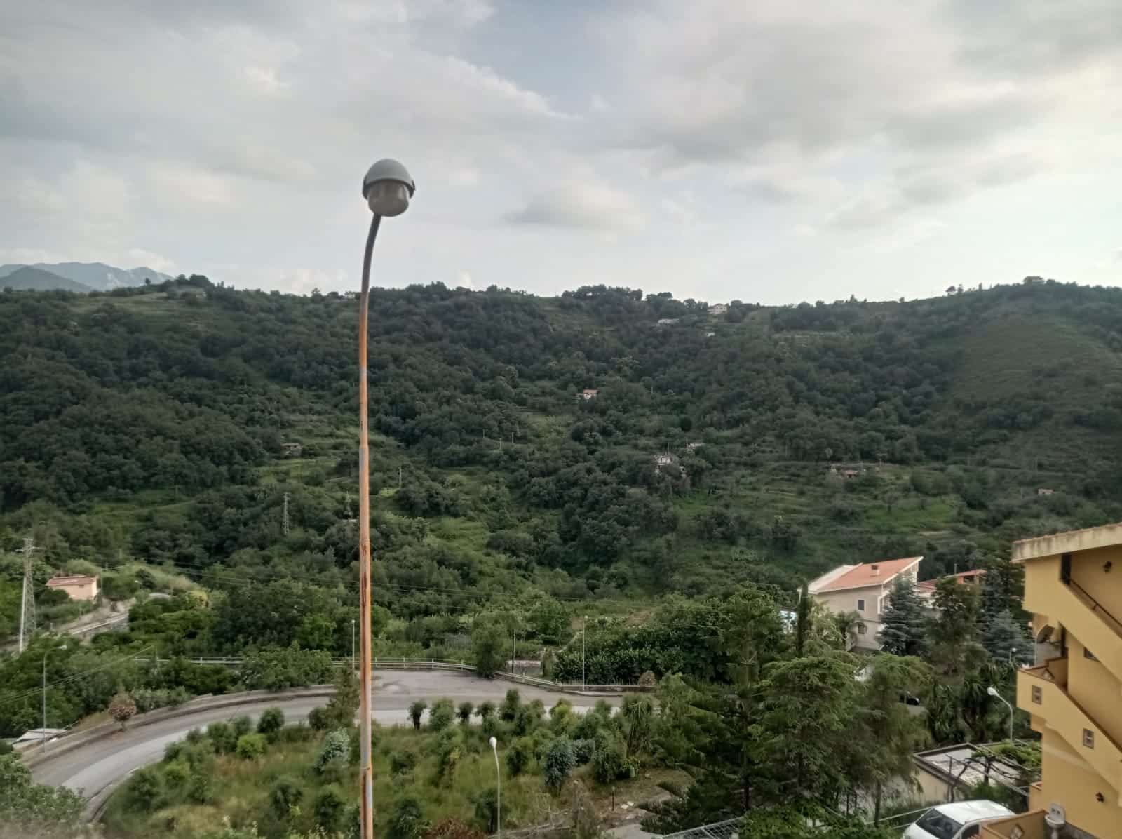 Finestra panoramica sulle colline verdi con strada curva, lampione e cielo nuvoloso, viste naturali per immobili, case e proprietà in vendita NextCasa.