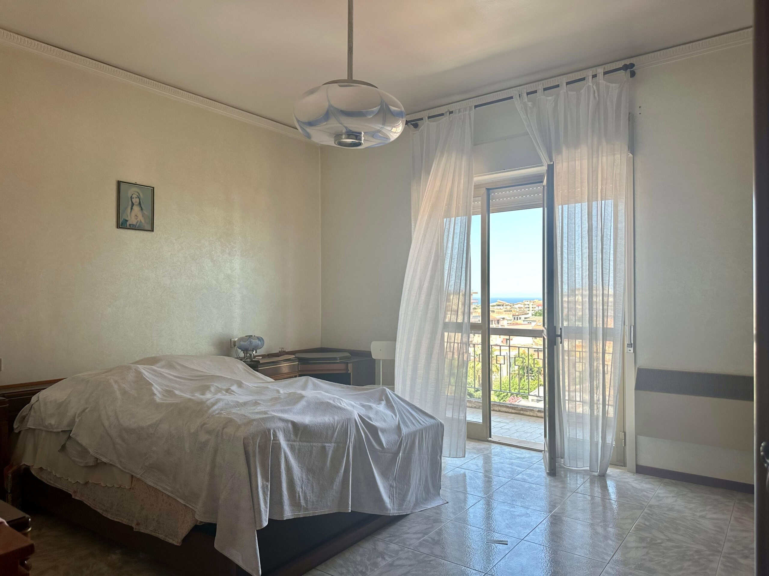 Camera matrimoniale con balcone e vista mare, arredi classici, luce naturale, tende leggere, ambiente tranquillo e accogliente, ideale per relax e riposo.