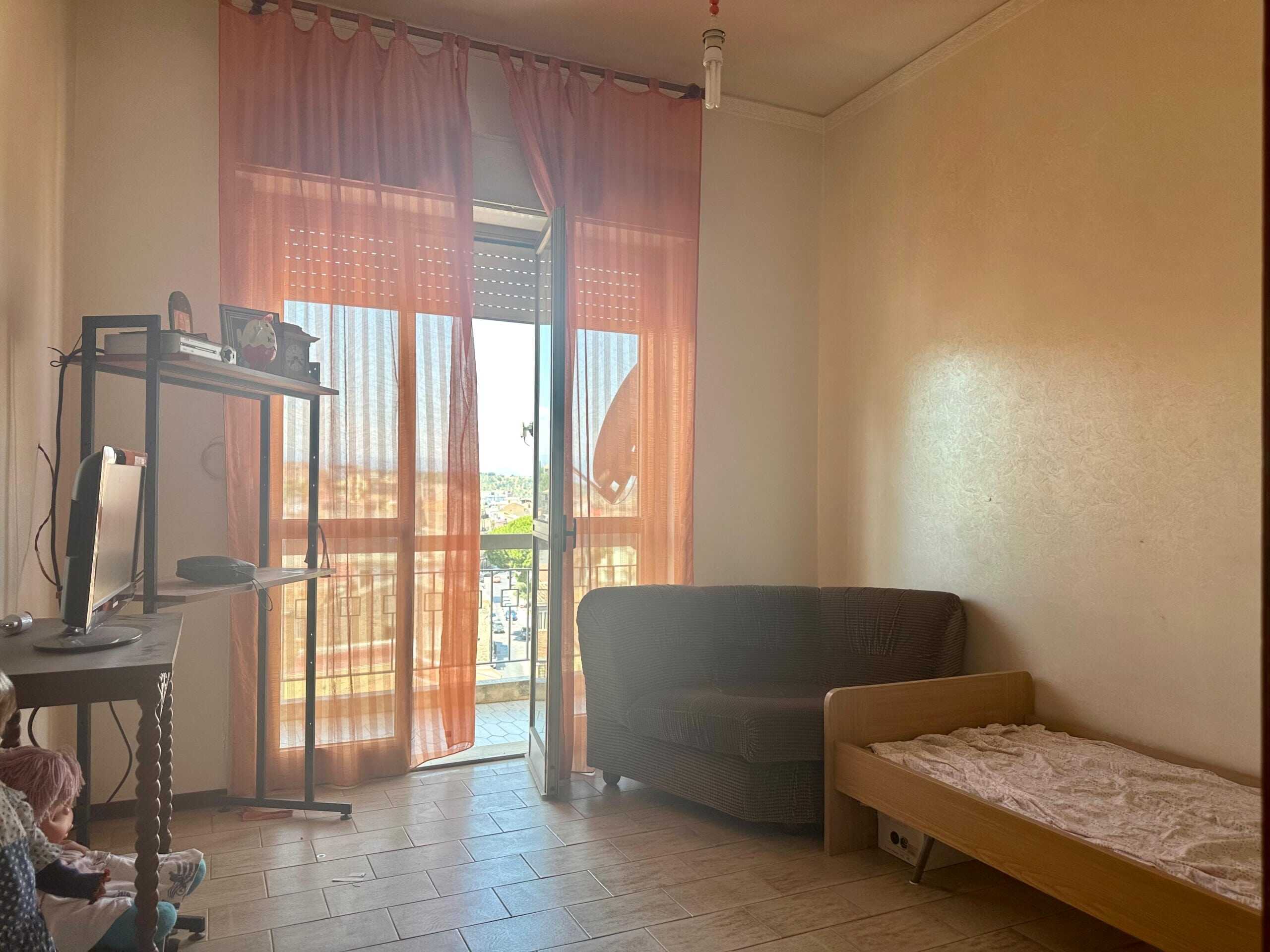 Balcone con vista cittadina e tende arancioni in soggiorno con divano, letto singolo in legno e piccolo televisore, ambiente luminoso e accogliente.