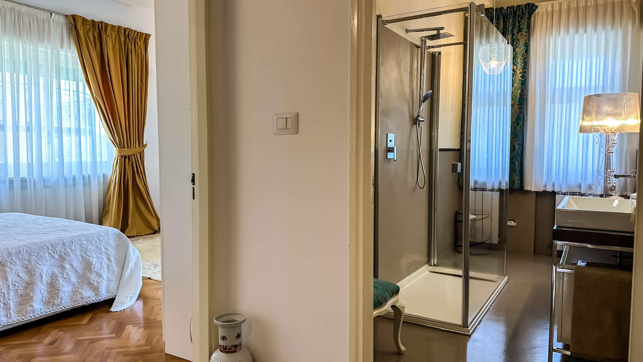 Arma da bagno con doccia e finestra, dettagli interni di un appartamento di lusso NextCasa, arredamento elegante, illuminazione calda, camera da letto visibile con tende dorate e biancheria bianca, stile sofisticato e confortevole.