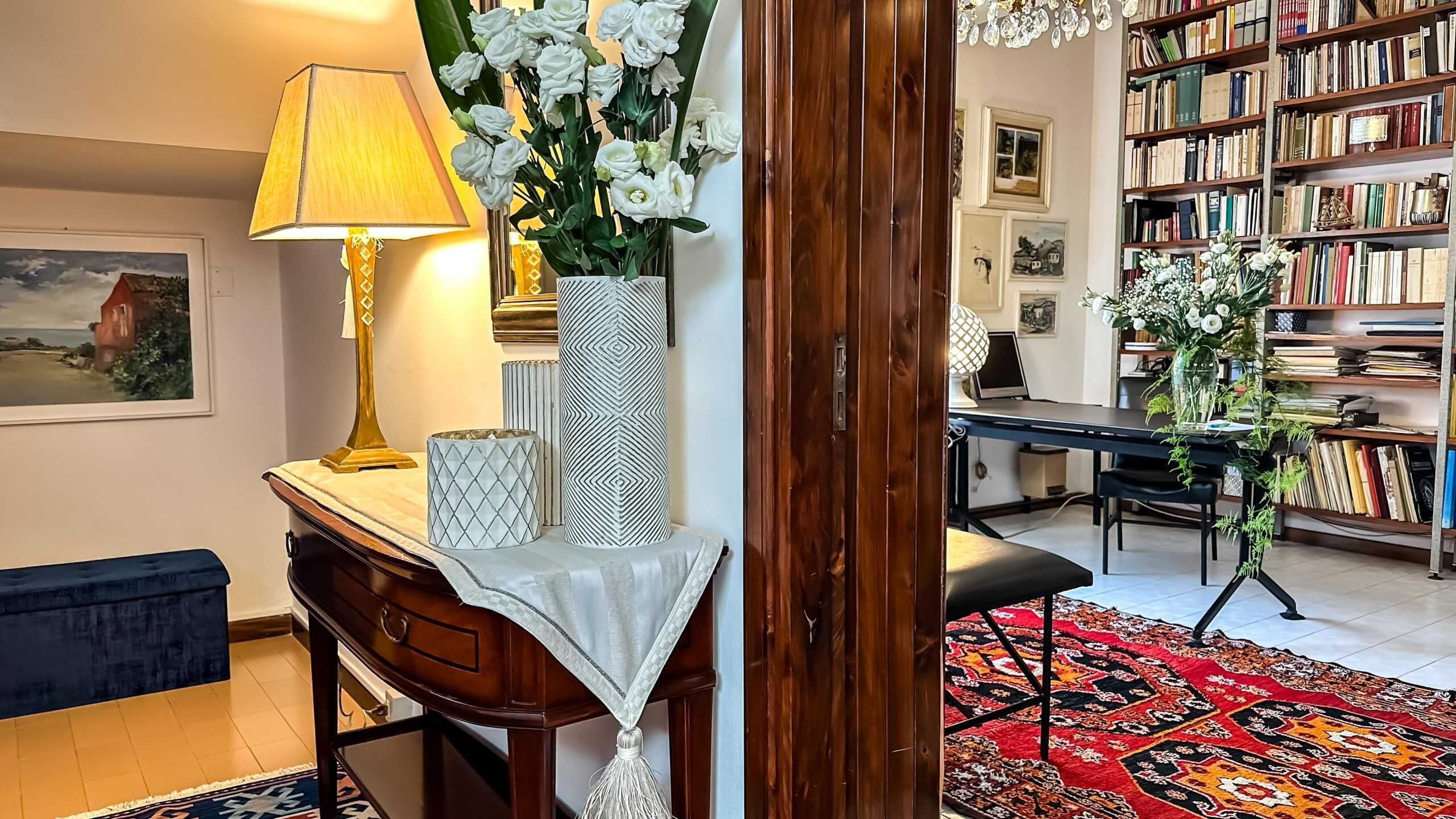 Vaso di fiori con bouquet bianco e dettagli decorativi in casa moderna, ambientazione con libreria, arredamento elegante e illuminazione calda, ideale per interni raffinati e accoglienti.