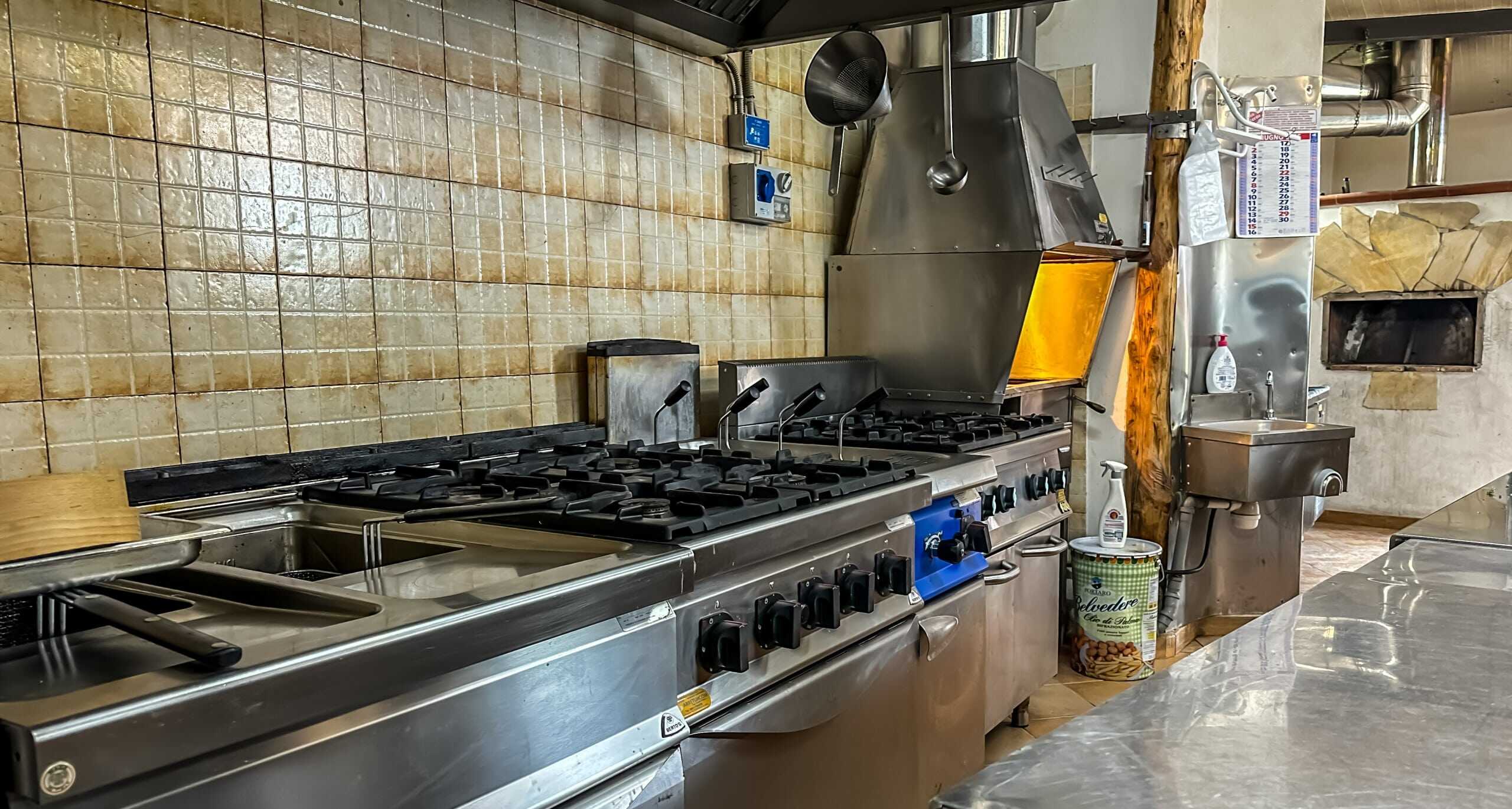 Cucina professionale con fornelli a gas, lavandino e attrezzature in acciaio inox, ambiente di ristorante NextCasa, utensili da cucina e pareti rivestite in piastrelle beige.
