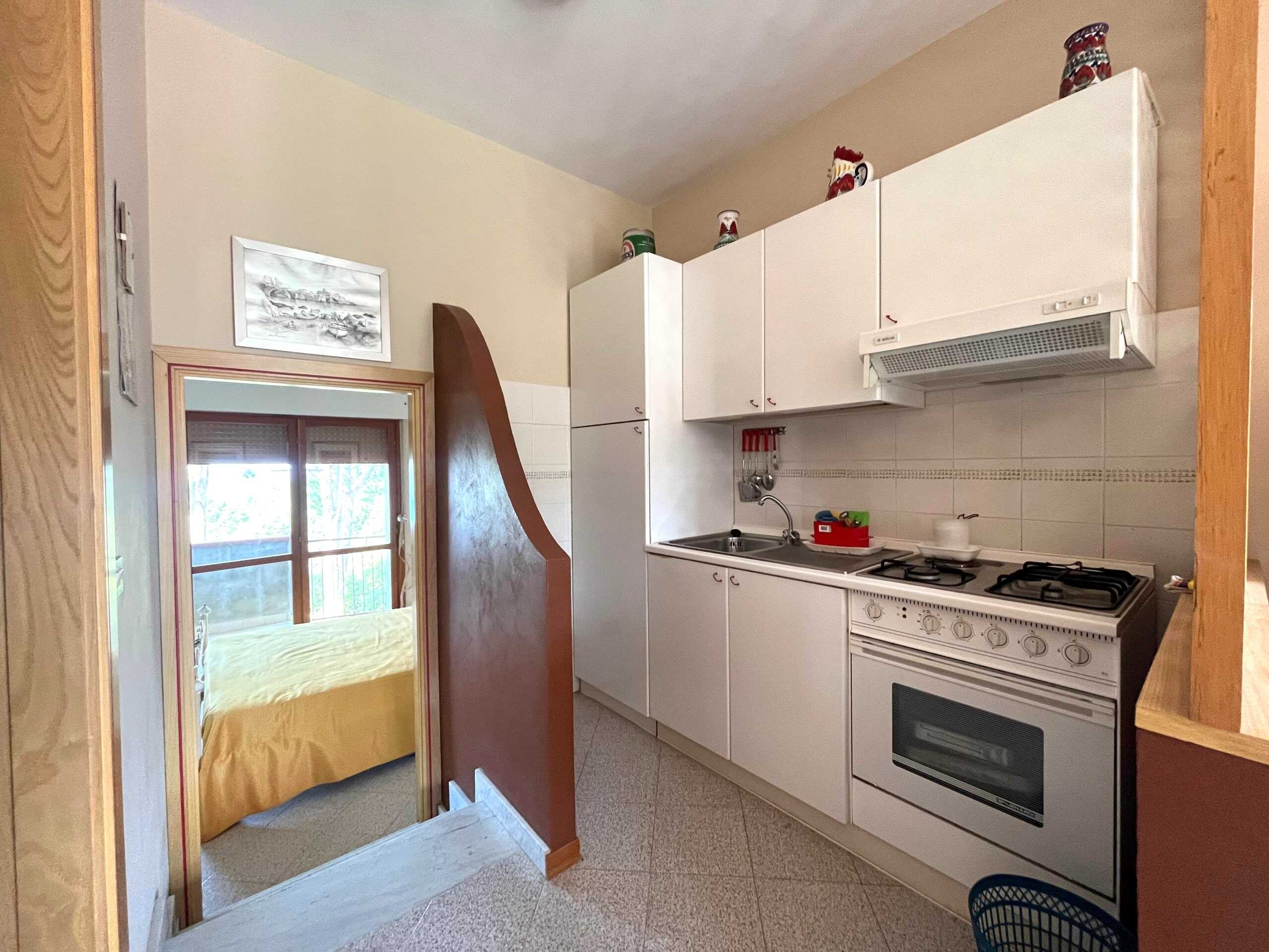 Cucina compatta con mobili bianchi e piano cottura a gas, ideale per appartamenti in affitto o case vacanza, con vista sulla camera da letto attraverso porta aperta, atmosfera funzionale e accogliente tutto cotidiano.