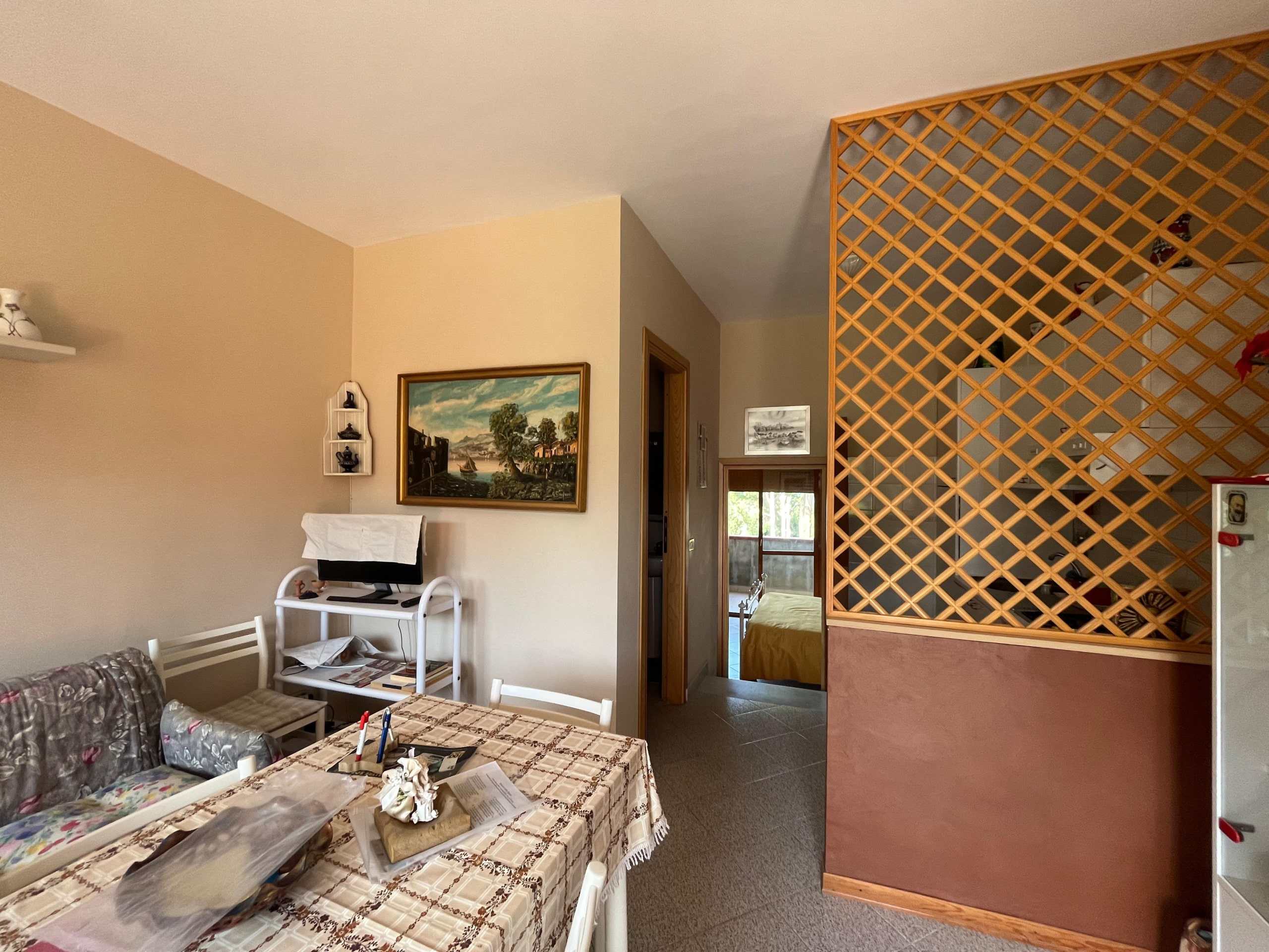Ampio soggiorno con pareti beige, tavolo da pranzo coperto da tovaglia a quadretti, divano, TV su supporto e decorazioni domestiche, separato da divisorio in legno a griglia, ambiente luminoso e accogliente.