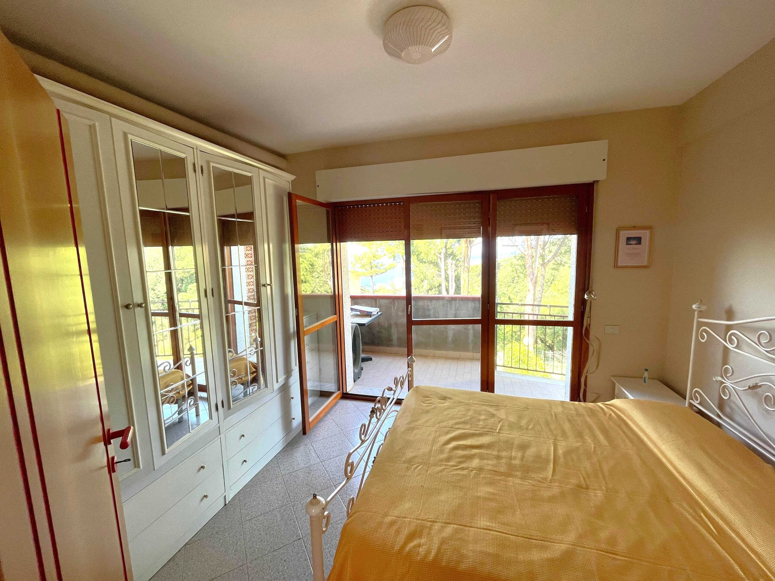 Vana camera da letto luminosa con grande balcone, armadio a doppio battente con specchi, letto con copriletto giallo, vista su giardino e natura, arredamento semplice e funzionale, accesso diretto all'esterno, luce naturale, ambiente tranquillo e accogliente.